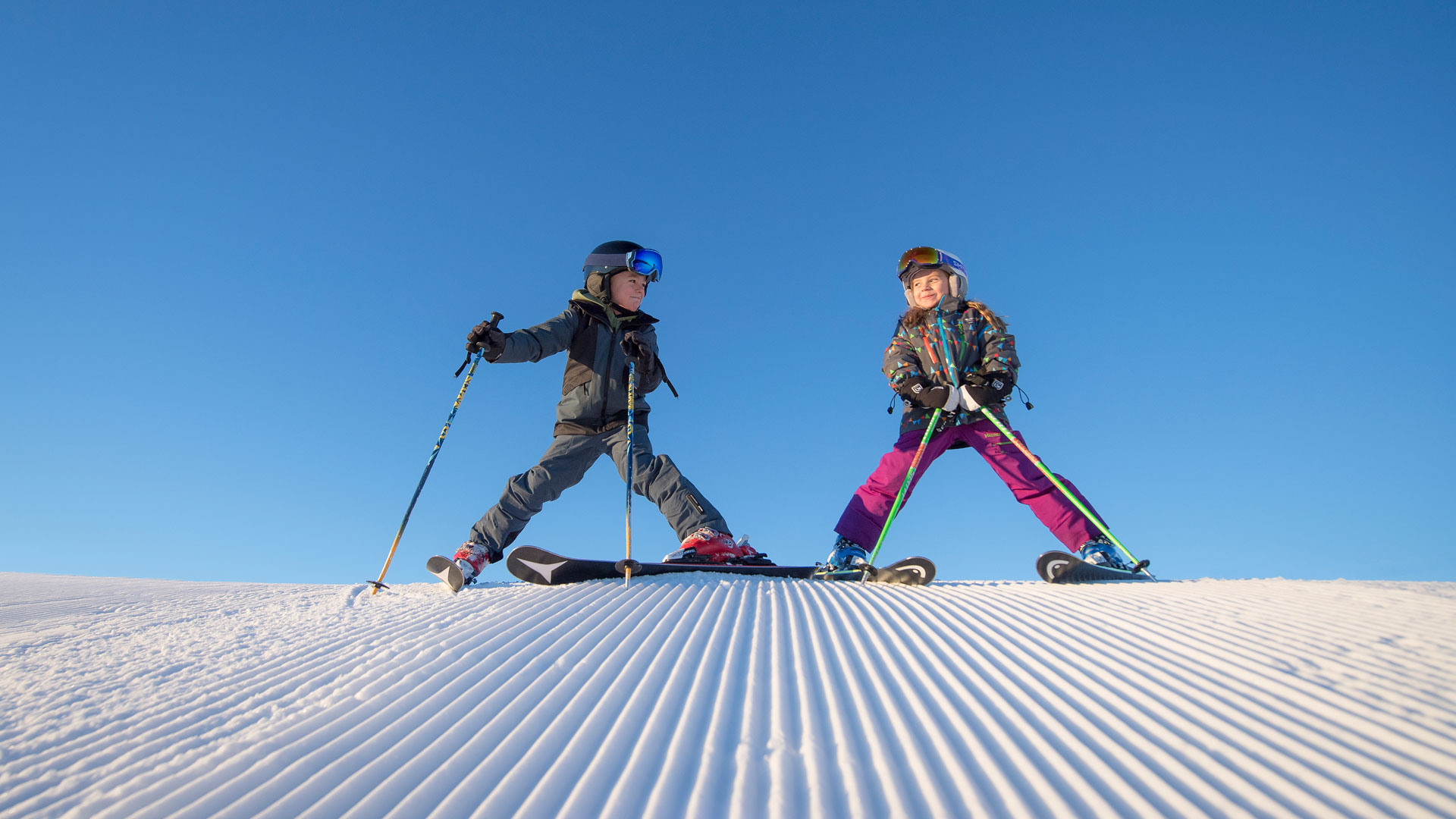 Två barn åker skidor på en solig vinterdag med klarblå himmel, iförda skidkläder och hjälm, njuter av nypistad snö