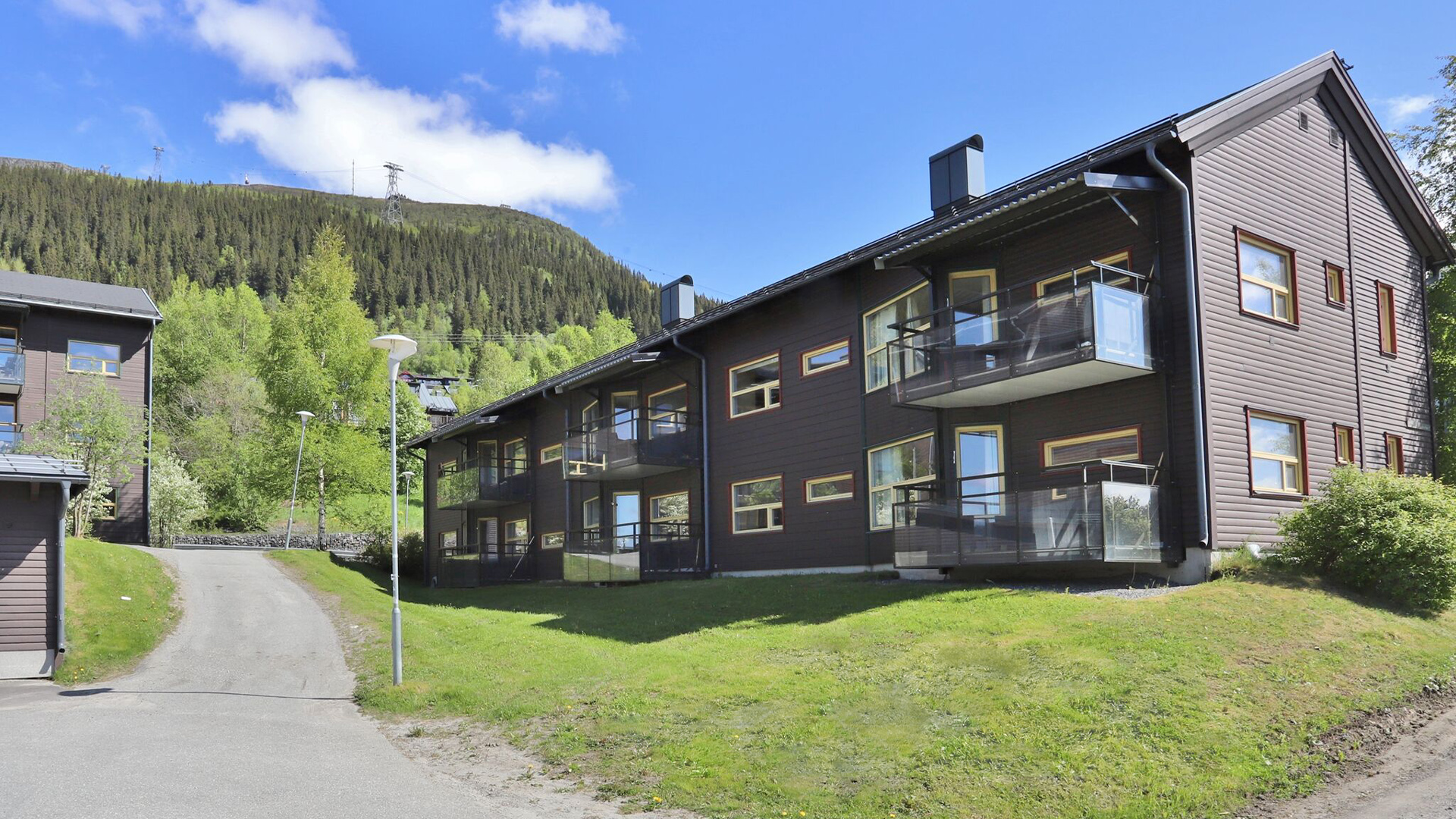 Wooden residential building with modern balconies, surrounded by lush nature and mountains in the background, under a clear blue sky