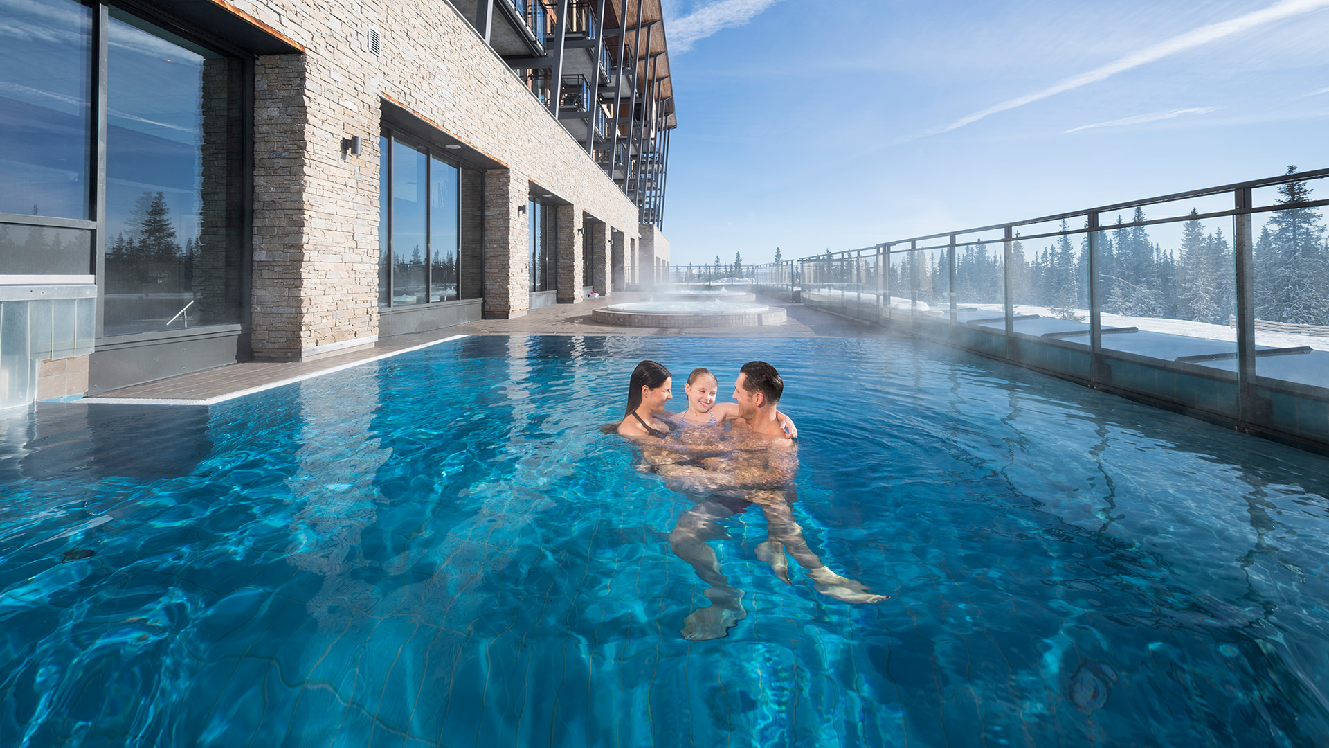 Personer svømmer i et oppvarmet utendørs basseng ved et hotell, med utsikt over snødekte trær og klar blå himmel