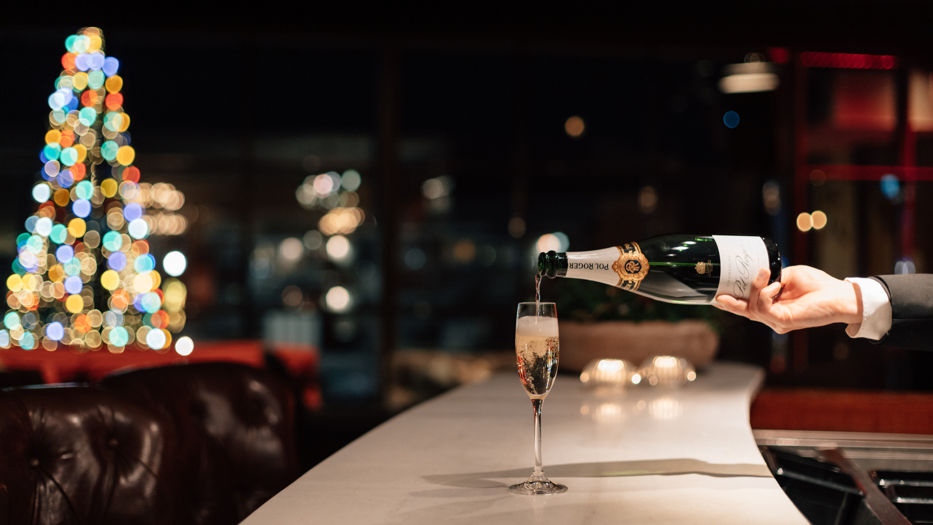 En person häller champagne i ett glas på en bardisk med en färgglad julgran i bakgrunden