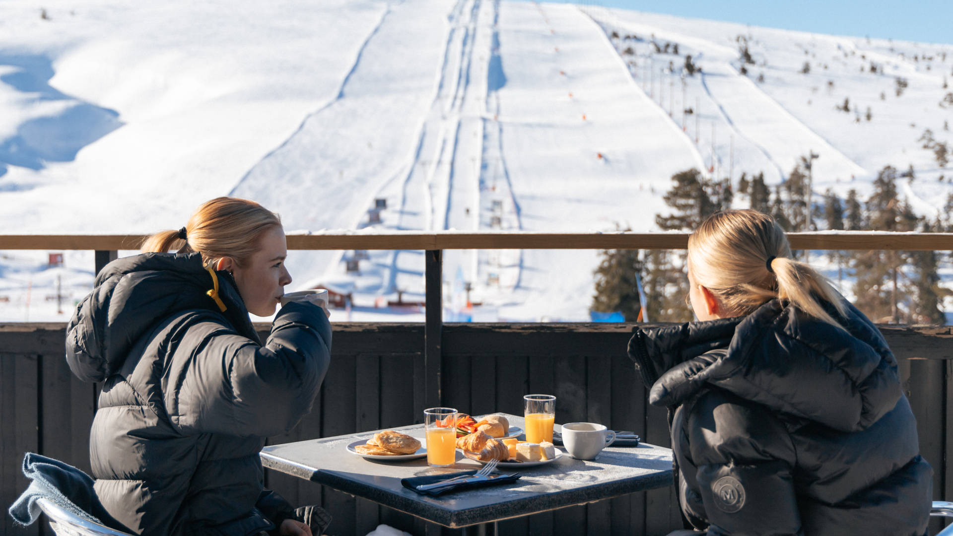 Två personer i vinterjackor sitter vid ett bord på en utomhusterrass med frukost och dryck, med utsikt över snötäckta skidbackar och blå himmel i bakgrunden