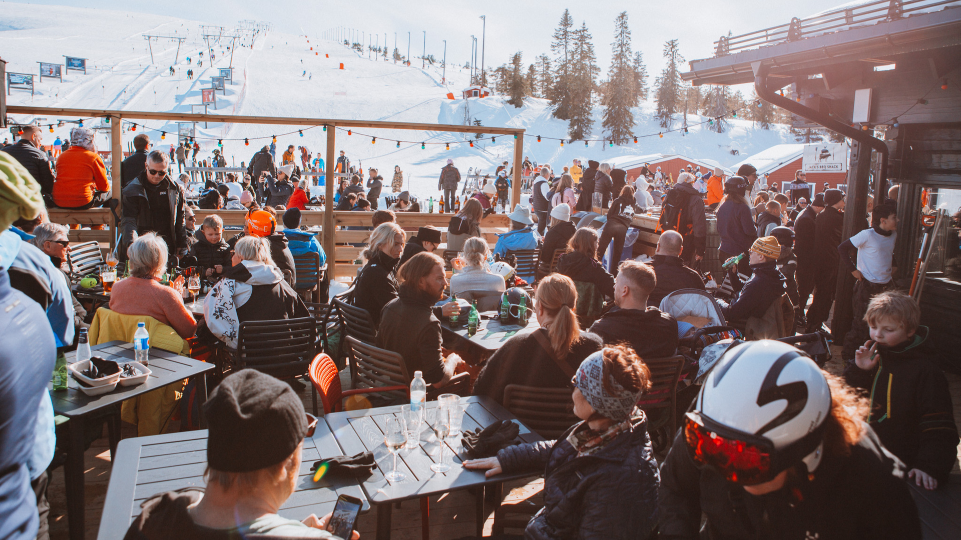 Uteservering på skidort i fjällen med många människor som njuter av soligt vinterväder, med snötäckta backar och träd i bakgrunden