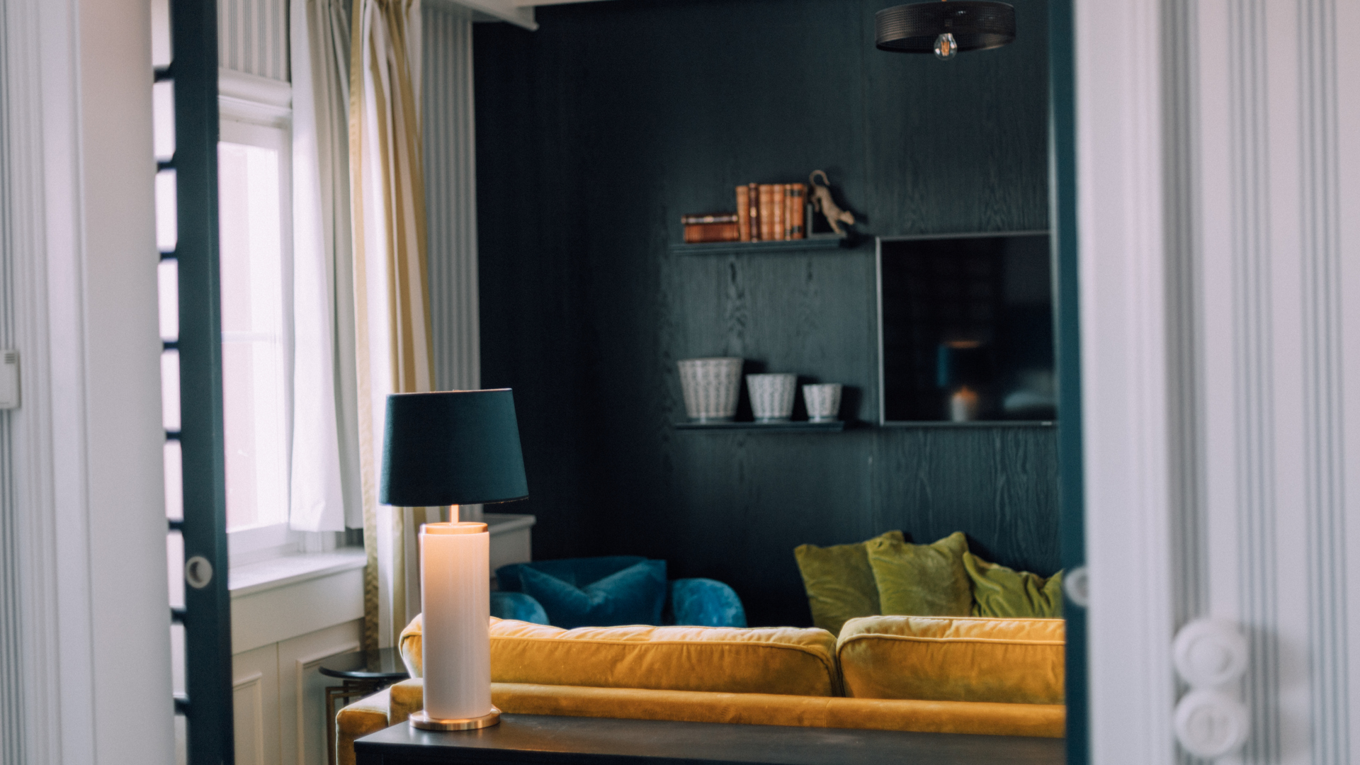 Modernt vardagsrum med mörka väggar, gul soffa och dekorativa kuddar, hylla med böcker och prydnad, samt bordslampa