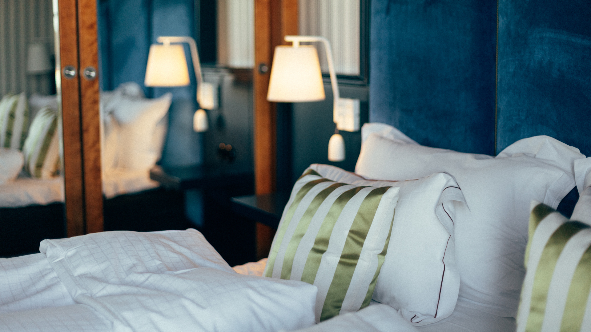 Lyxig hotellsäng med vita sängkläder och grönrandiga kuddar, belyst av eleganta sänglampor mot en blå sänggavel