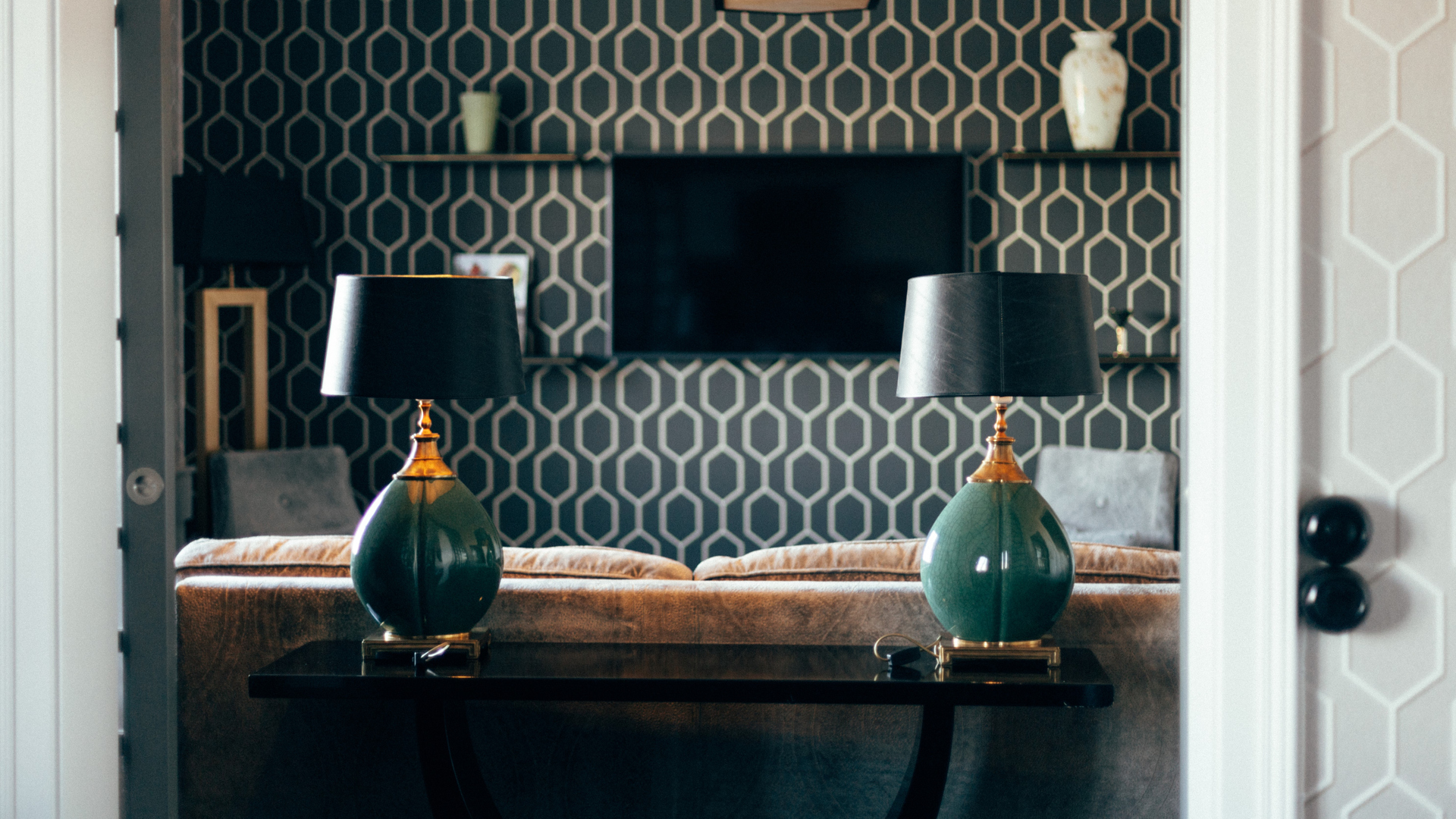 Elegant vardagsrum med två gröna bordslampor på ett träbord framför en mönstrad vägg med hyllor och en TV