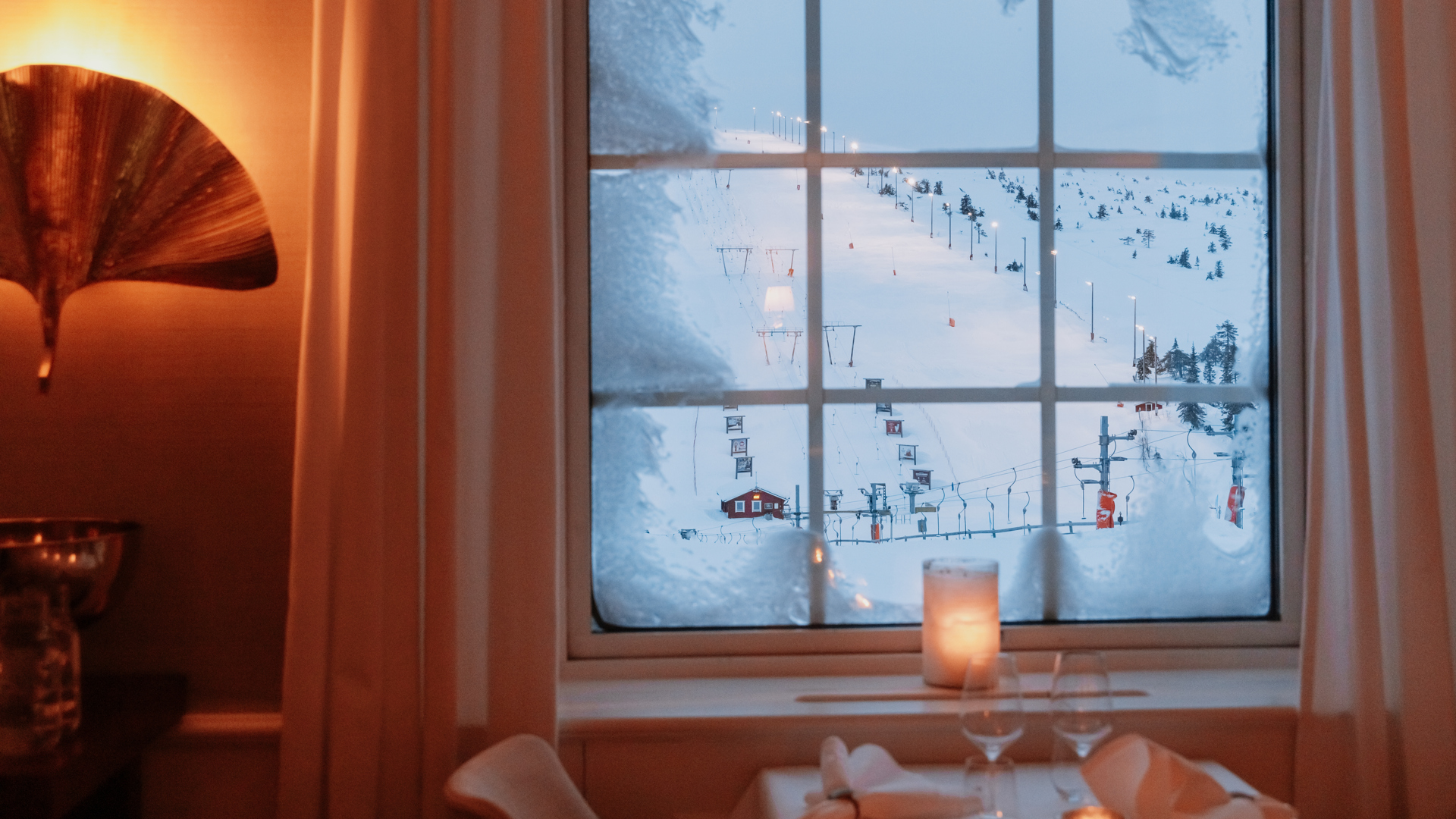 Vy från fönster över snöig skidbacke med tända ljus och dukat bord inomhus