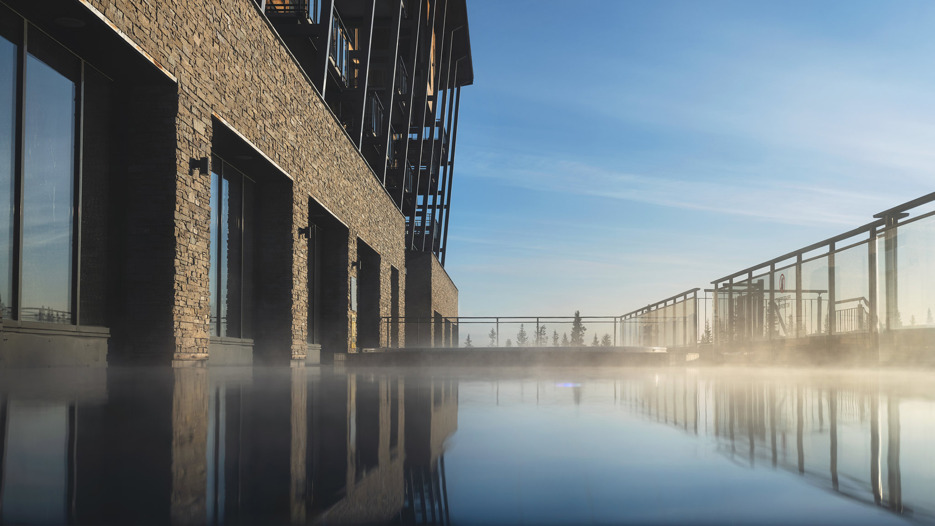 Modern byggnad med glas och stenfasad vid en ångande infinitypool under klarblå himmel