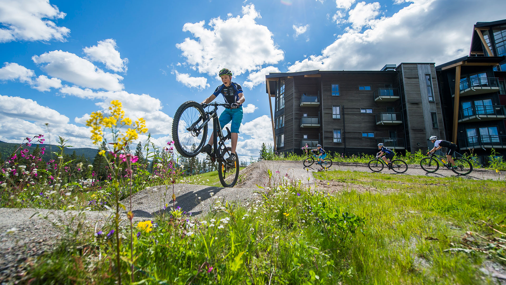 Person som cyklar mountainbike i en naturskön miljö med flera cyklister och moderna byggnader i bakgrunden under en solig dag