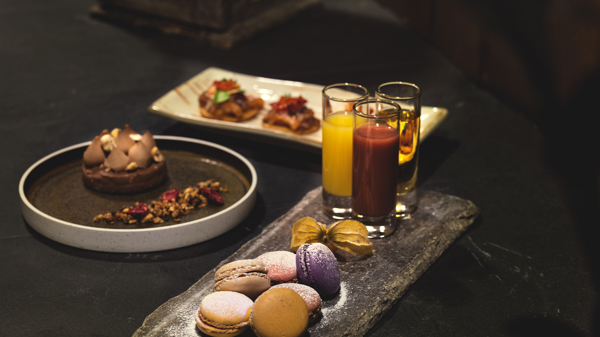 En elegant dessertoppsats med makroner, en sjokoladekake, små matbiter og tre fargerike juiceglass på en svart bakgrunn