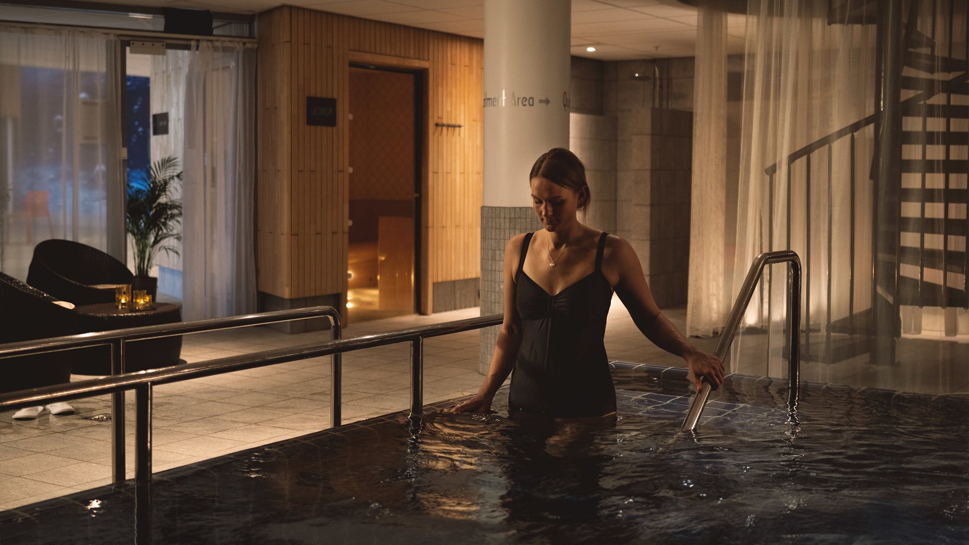 Kvinna i svart baddräkt kliver i en varm pool på ett spa, omgivet av påkostad inredning och dämpad belysning