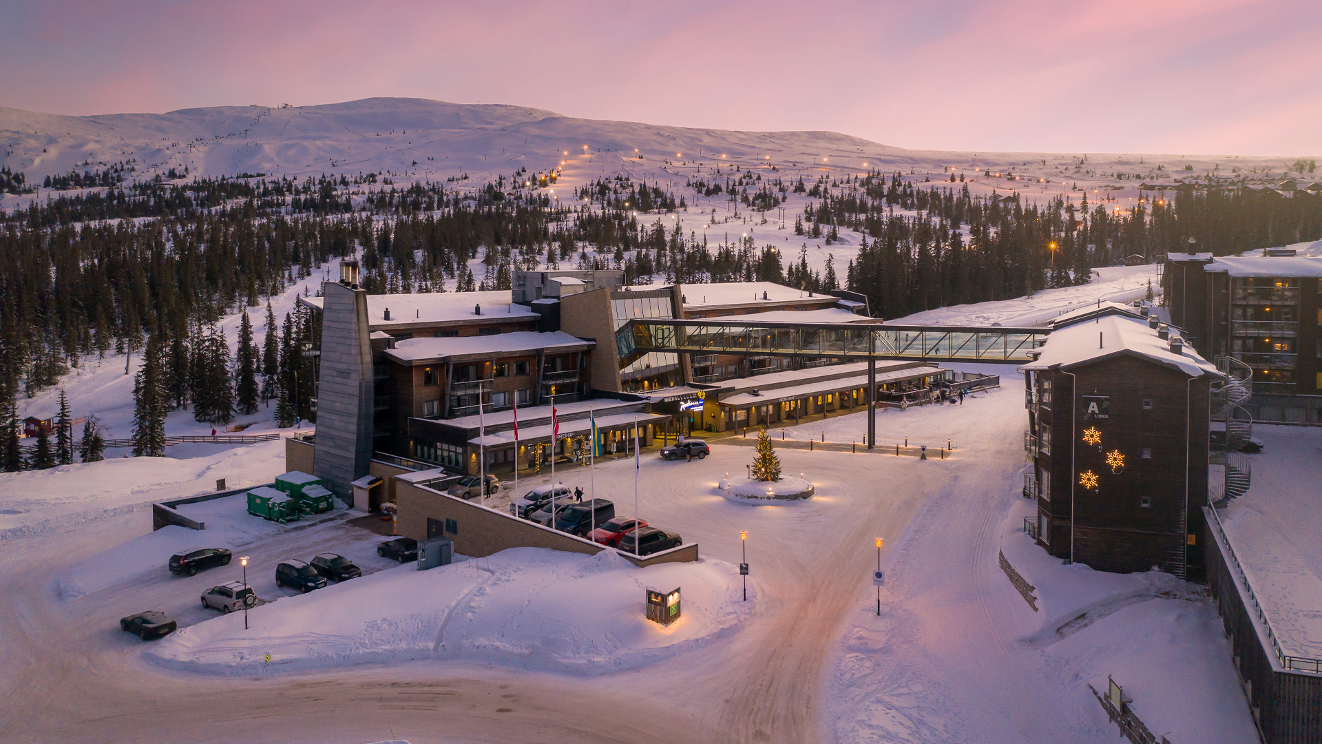 Winterliches Resort in verschneiter Berglandschaft bei Sonnenuntergang, mit modernen Gebäuden, Brücke und dekorativen Lichtern