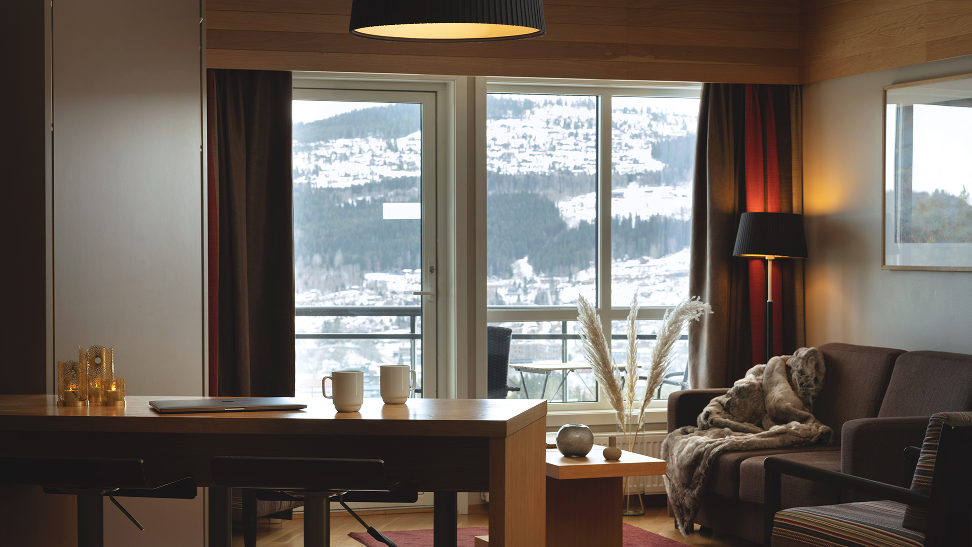 Modernt vardagsrum med snöig bergsutsikt genom stora fönster. Inredningen inkluderar en soffa, bord med kaffekoppar och dekorativa föremål, samt belysning för en mysig atmosfär