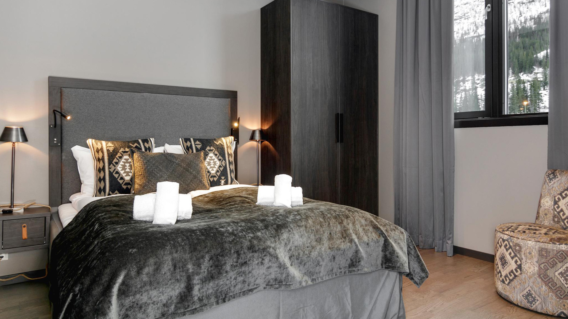 Moderne hotellrom med dobbeltseng, dekorative puter og håndklær, innrammet av tepper og lampeskinn, med utsikt mot snødekte fjell gjennom vinduet
