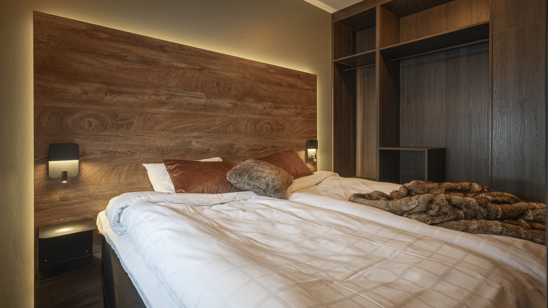 Sovrum med modern inredning, mörk trähuvudgavel, vita sängkläder och bruna kuddar, samt en öppen garderob i ett lyxigt hotellrum