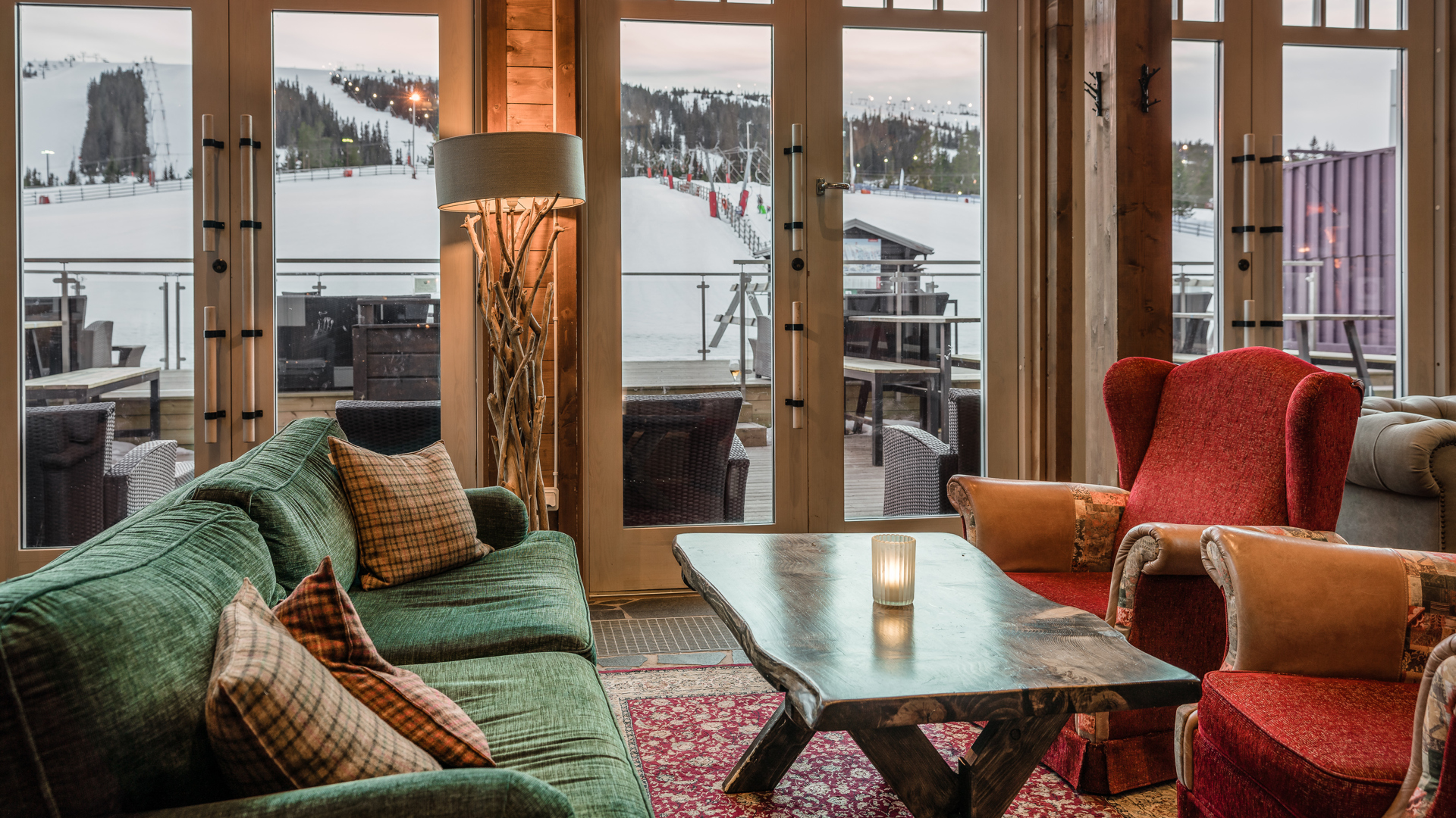 Loungeområde med bekväma soffor och stolar vid fönster som vetter mot snötäckta berg och en skidbacke, med ett mysigt ljus placerat på ett rustikt träbord