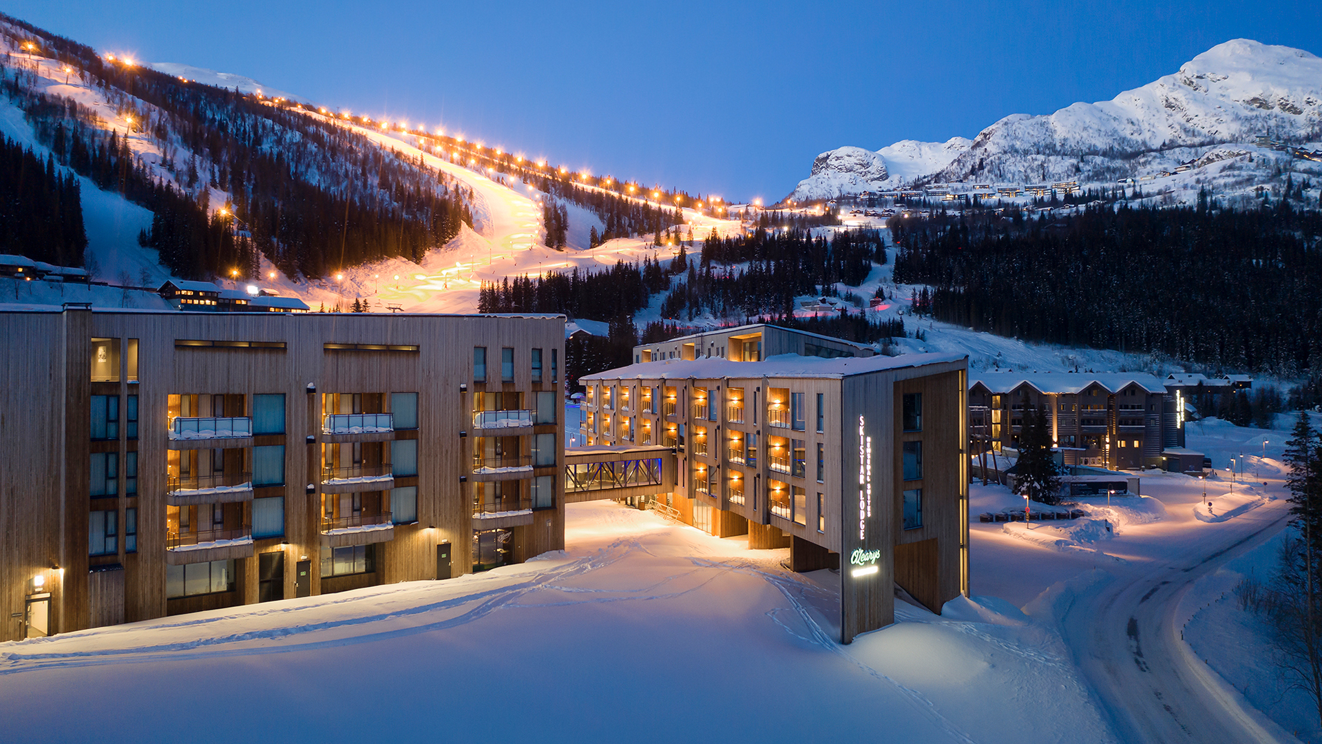 Moderna hotellbyggnader vid en snötäckt skidort med upplysta backar och majestätiska berg i bakgrunden under skymningen i Hemsedal