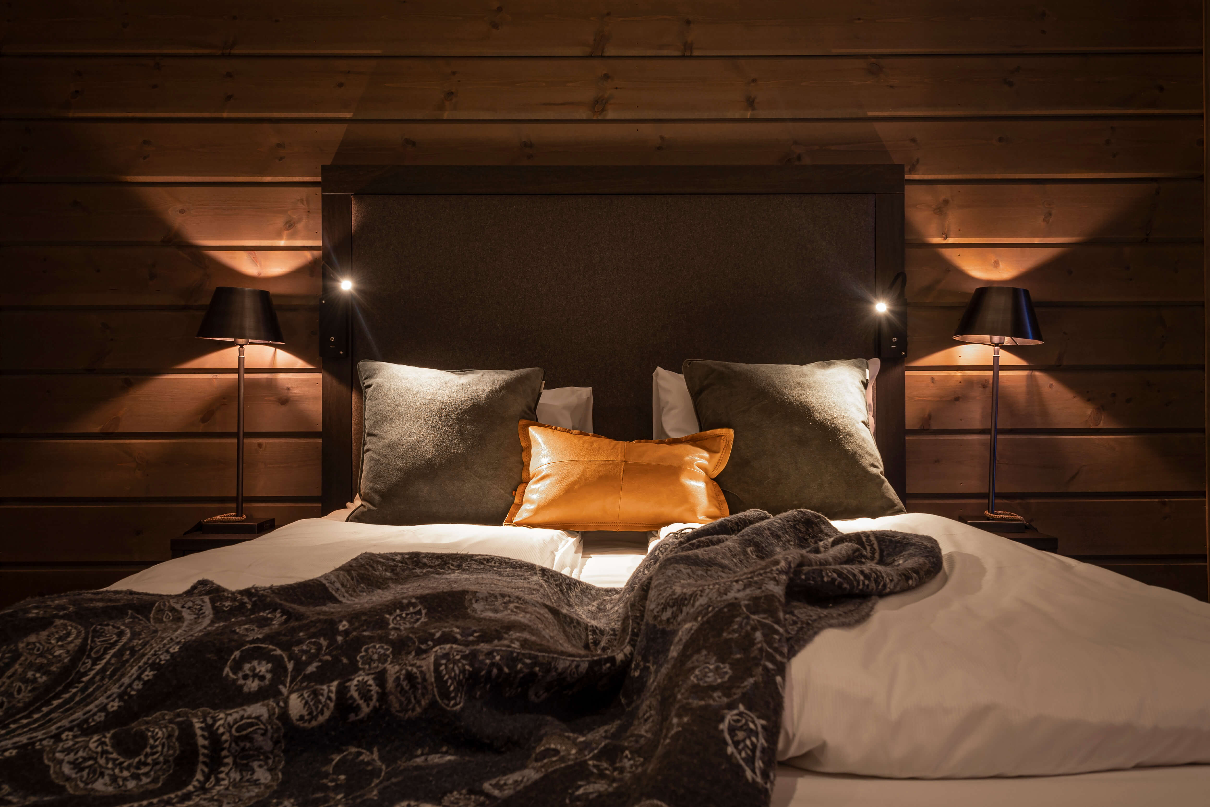 Säng med bruna träväggar, stilrena sänglampor och mysiga kuddar i naturskalor, inklusive en brun läderkudde, ger en varm och inbjudande atmosfär