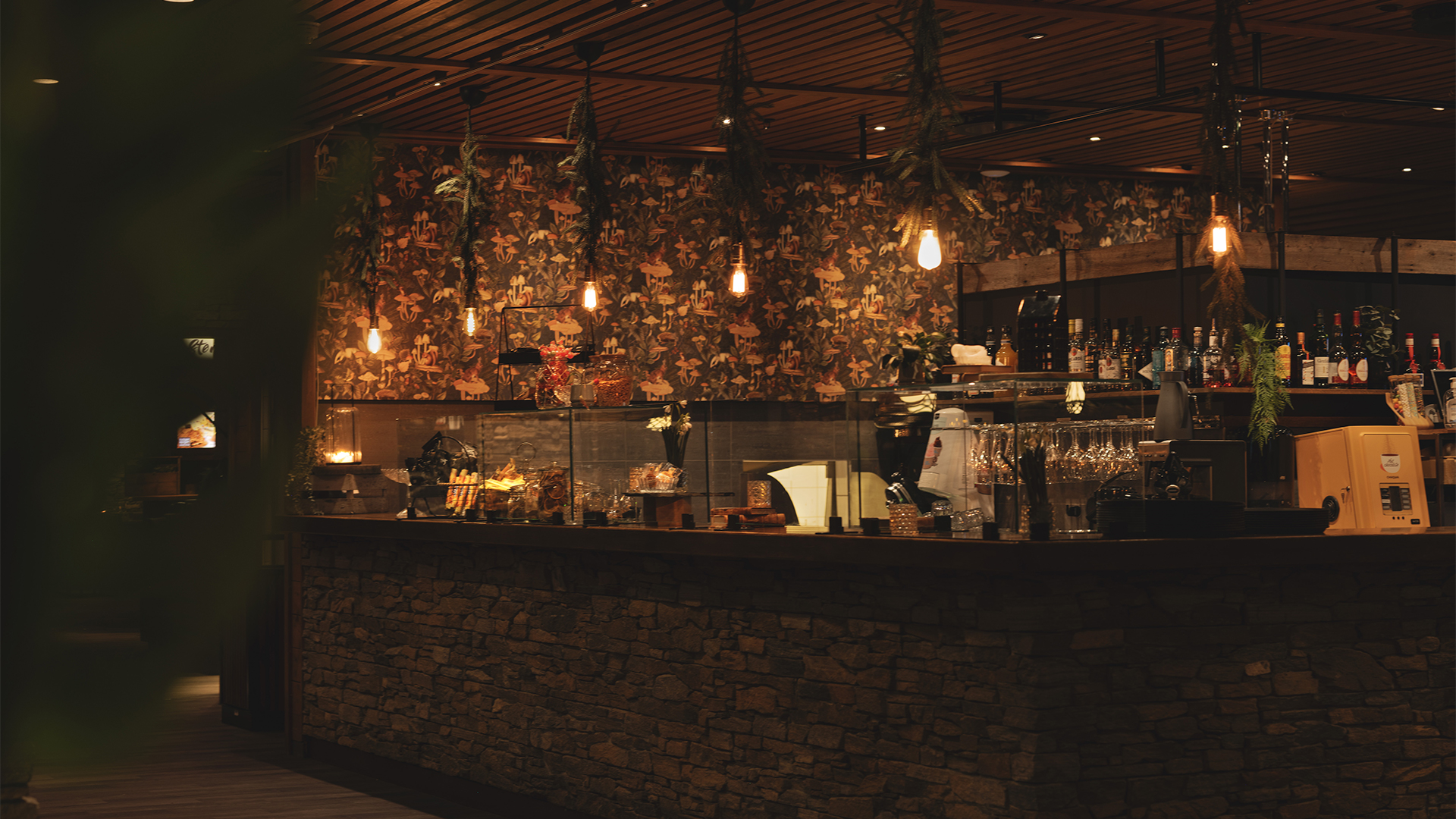 Rustik barinteriör med dämpad belysning och mönstrad tapet i bakgrunden. Flaskor och glas står prydligt uppradade på bänken, med hängande lampor ovanför