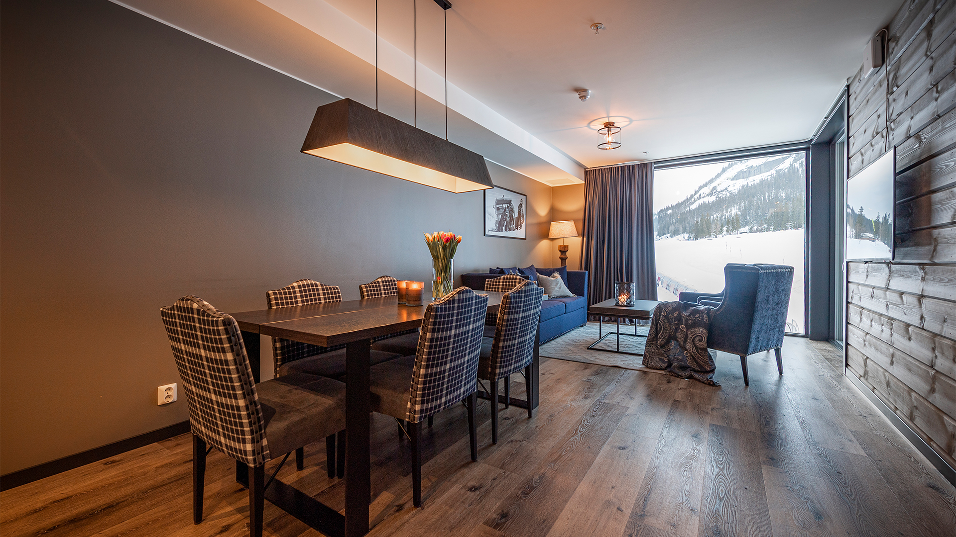 Modernt vardagsrum med plädklädda stolar kring ett matbord, bredvid en soffa och fåtölj i en stuga med utsikt över snöiga berg