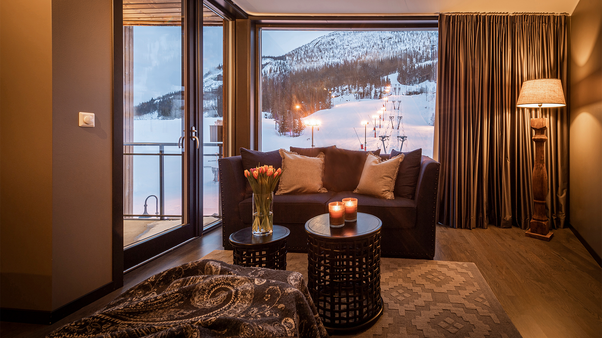 Vardagsrum med utsikt över snöigt landskap, mysig inredning med soffa, tända ljus och blommor i vas vid fönster