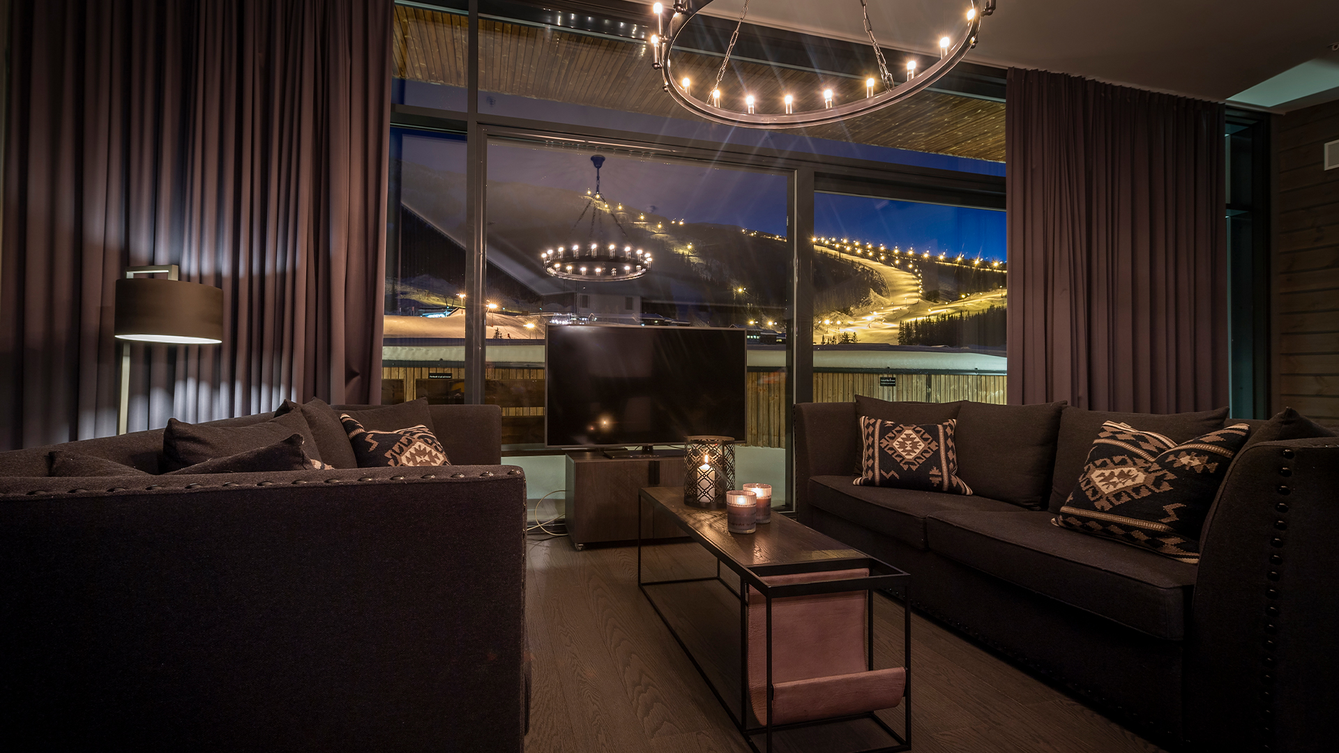 Modern och stilfullt vardagsrum med mörka soffor, kuddar med mönster och ljuskrona. Stora fönster visar en snötäckt skidbacke upplyst på kvällen utanför