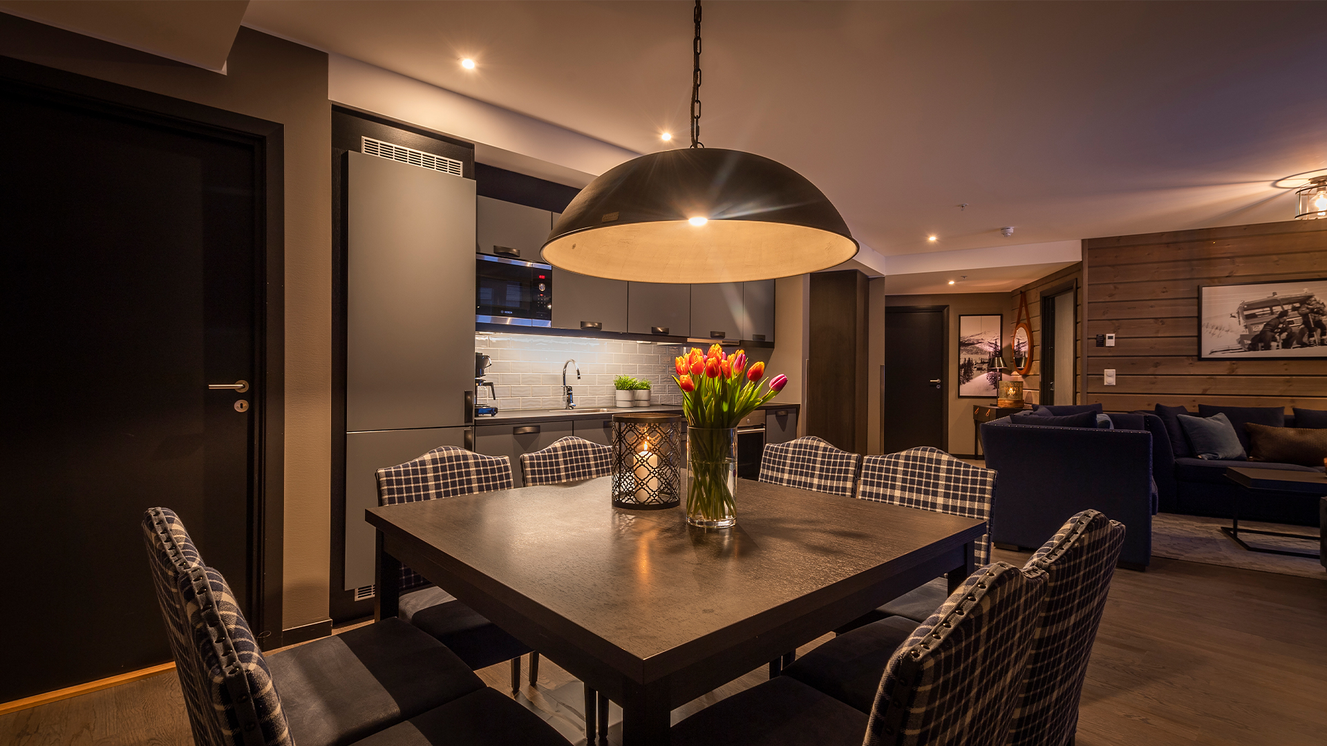Modernt kök med mörkt trämöblemang, en vas med färgglada tulpaner på bordet och en stor taklampa. Bakom syns ett designat vardagsrum och en stilren inredning i jordnära toner