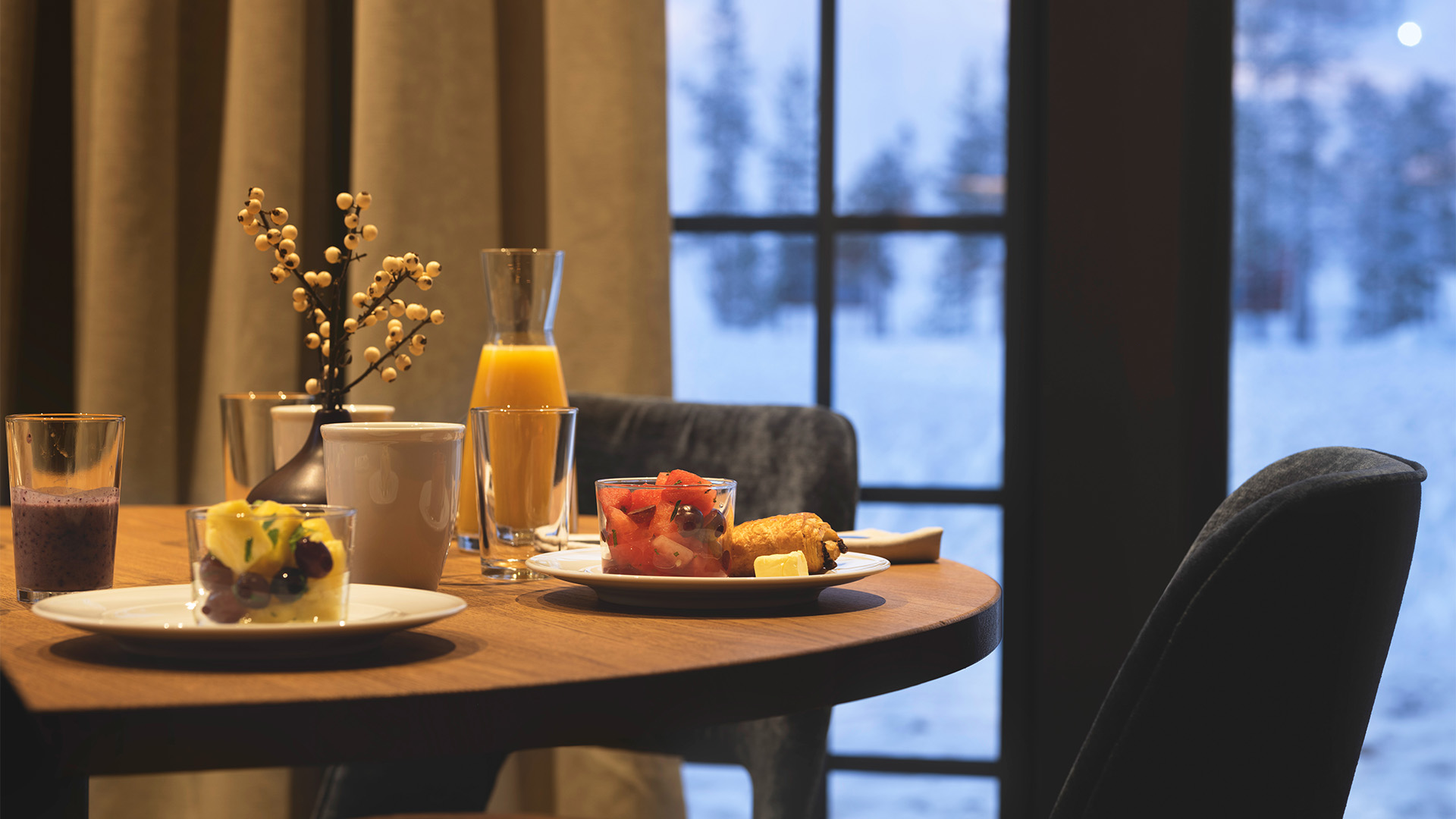 Frukostbord med olika maträtter inklusive frukt, en croissant och juice, med utsikt över ett snölandskap genom ett fönster