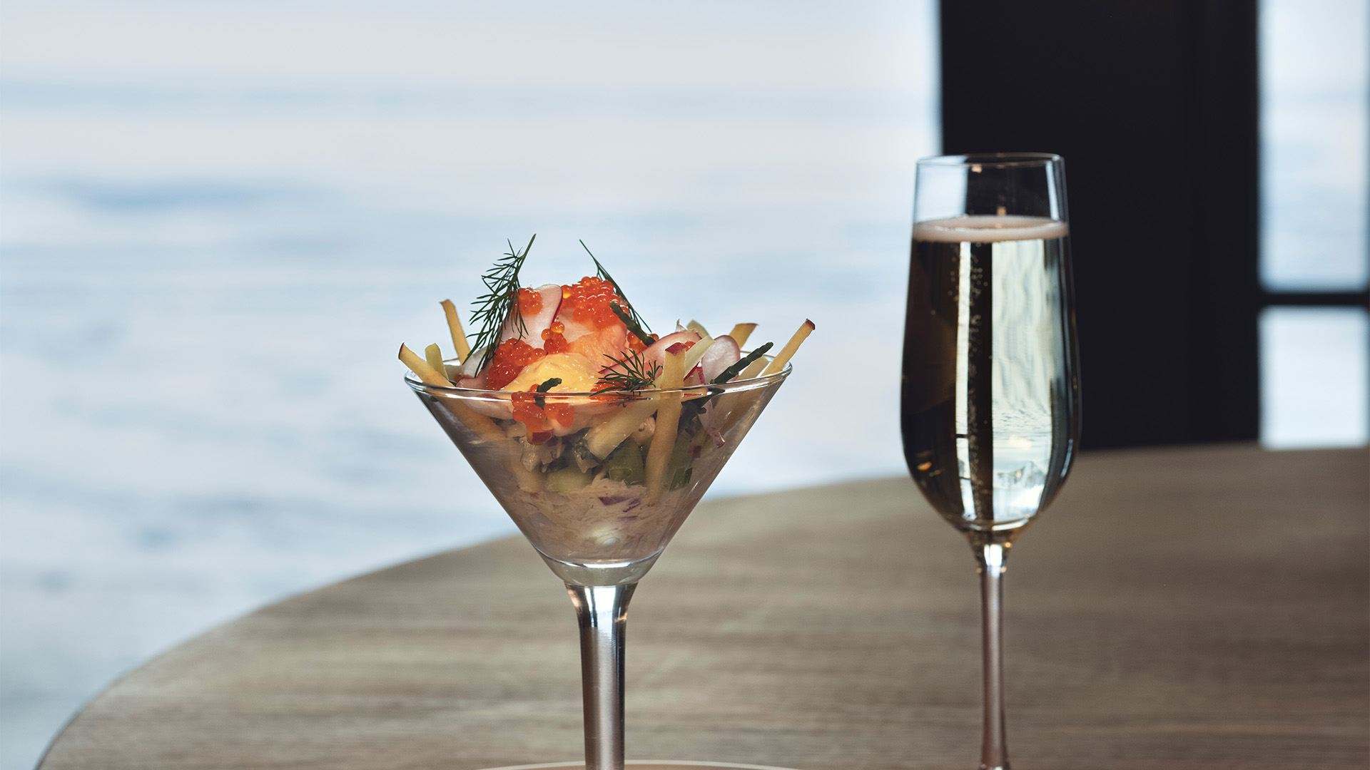 Cocktailglas med skaldjurscocktail dekorerad med rom och dill, bredvid ett glas mousserande vin på ett bord med havsutsikt