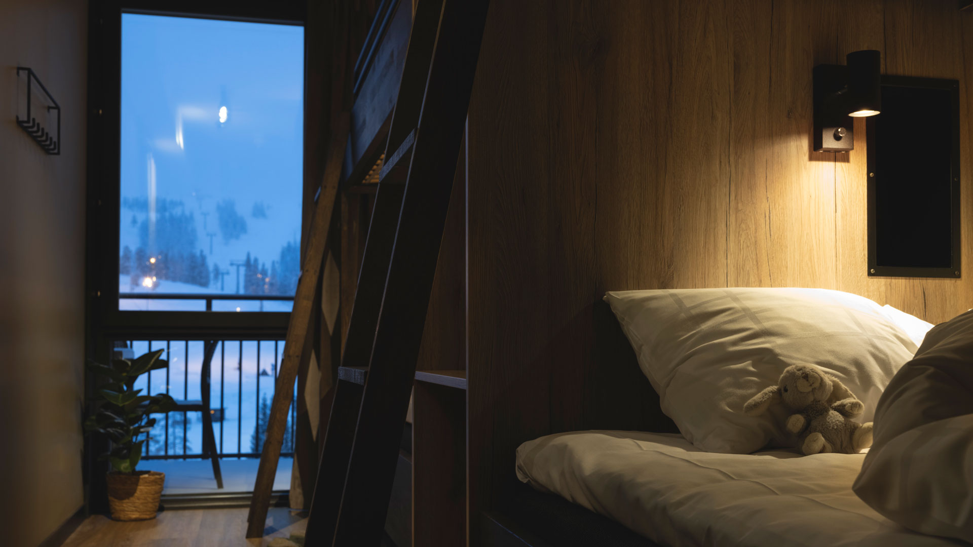 Mysig sovplats i träinspirerat rum med fönsterutsikt över snölandskap, växt i hörnet och en nalle på kudden