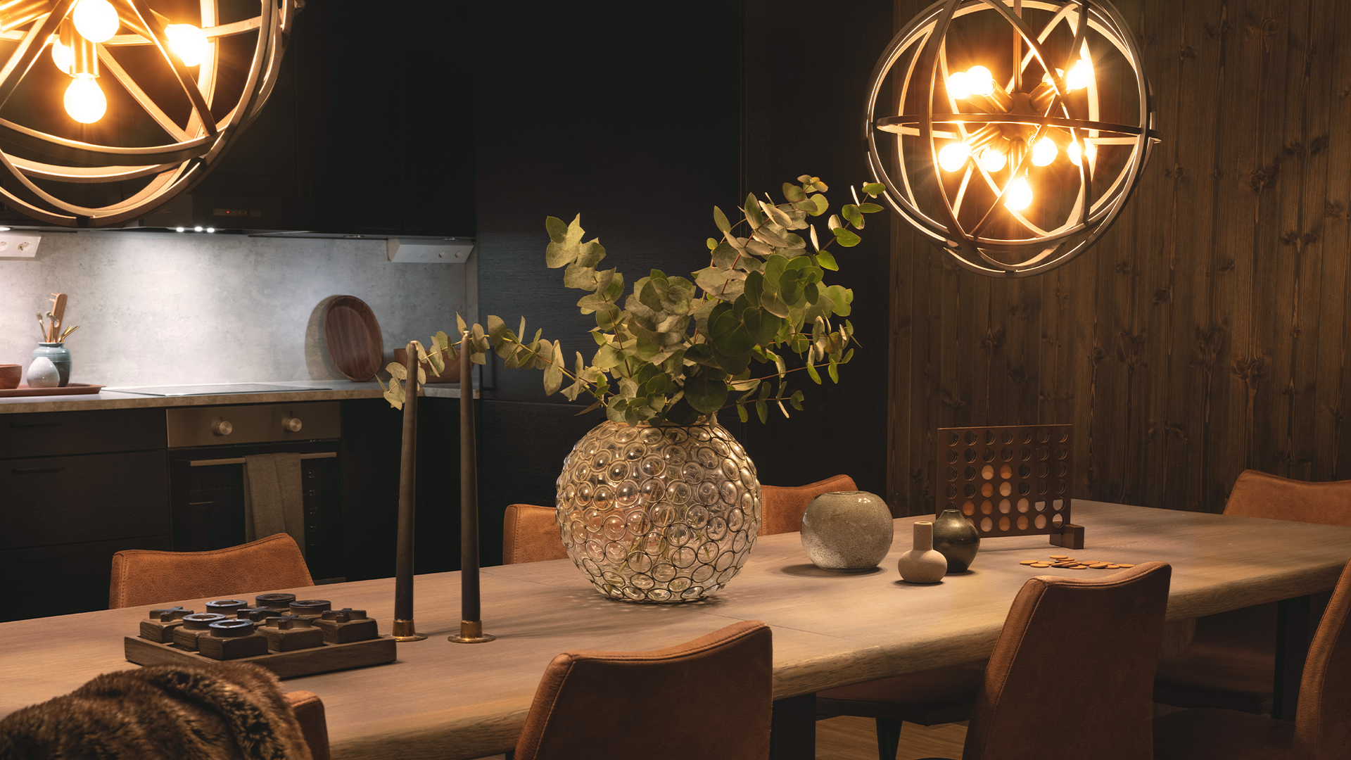 Modernt matrum med elegant belysning, stor glasvas med gröna kvistar som dekoration på träbord, omgiven av bekväma stolar i läder