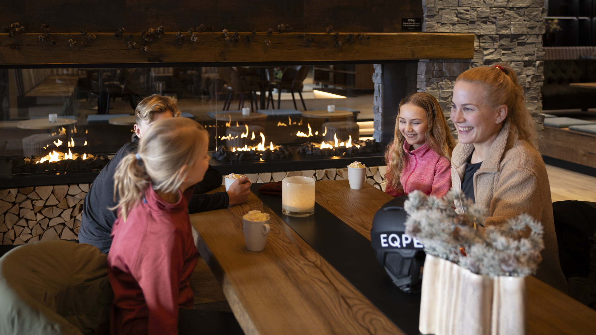 Fyra personer njuter av varma drycker framför en öppen spis i en mysig, rustik restaurangmiljö