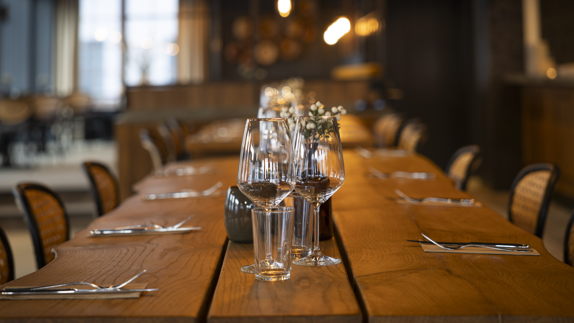 Dukat träbord i restaurangmiljö med vinglas och bestick, stämningsfull belysning i bakgrunden