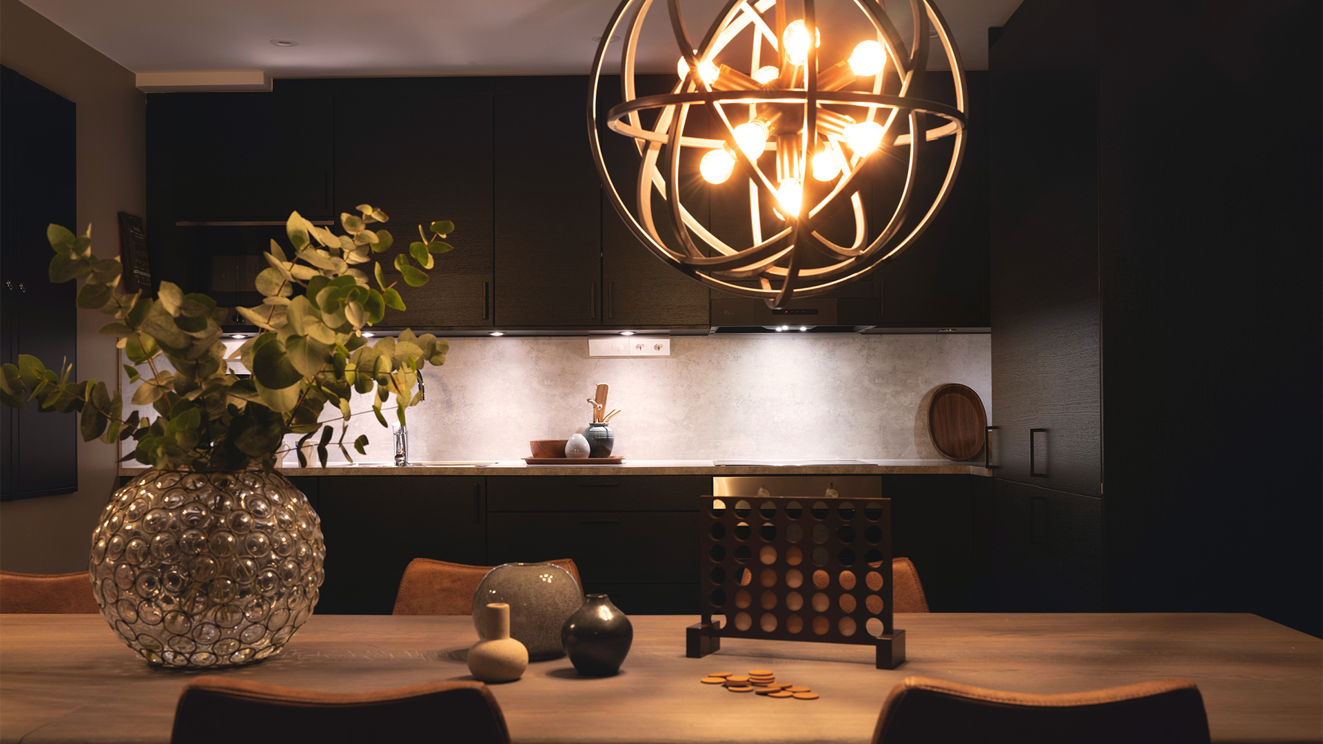 Modernt kök med stilren inredning, mörka skåp, och en dekorativ, rund lampa ovanför bordet. Bordet är dekorerat med vaser och en växt i en glasvas