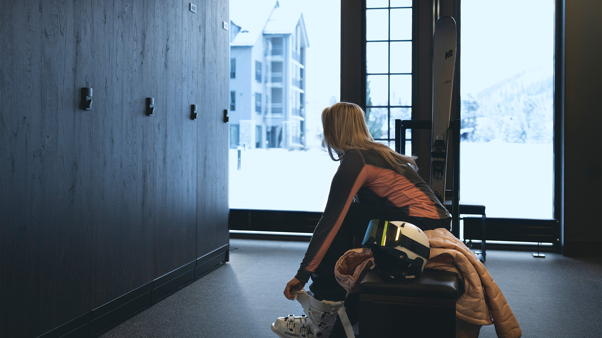 Kvinna sittande i skidstuga, justerar skidpjäxor, med hjälm och skidor bredvid