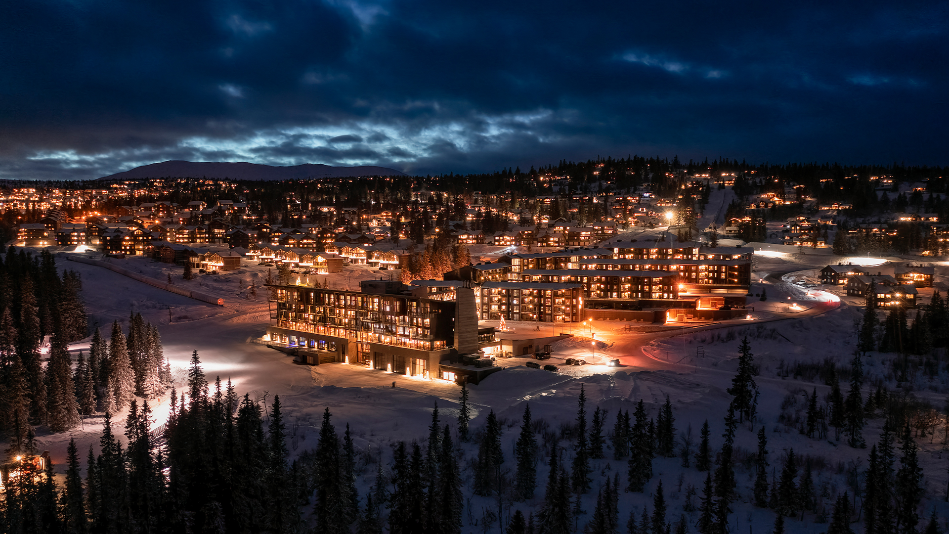 Snöig bergssemesterby med upplysta träbyggnader i skymningen, omgiven av skog