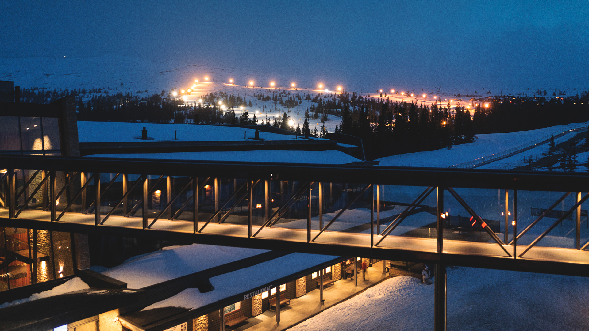 Nattvy av skidort i fjällen med snötäckta backar och lysande lampor. En inglasad gångväg löper över byggnader i förgrunden och skapar en modern kontrast mot den natursköna omgivningen