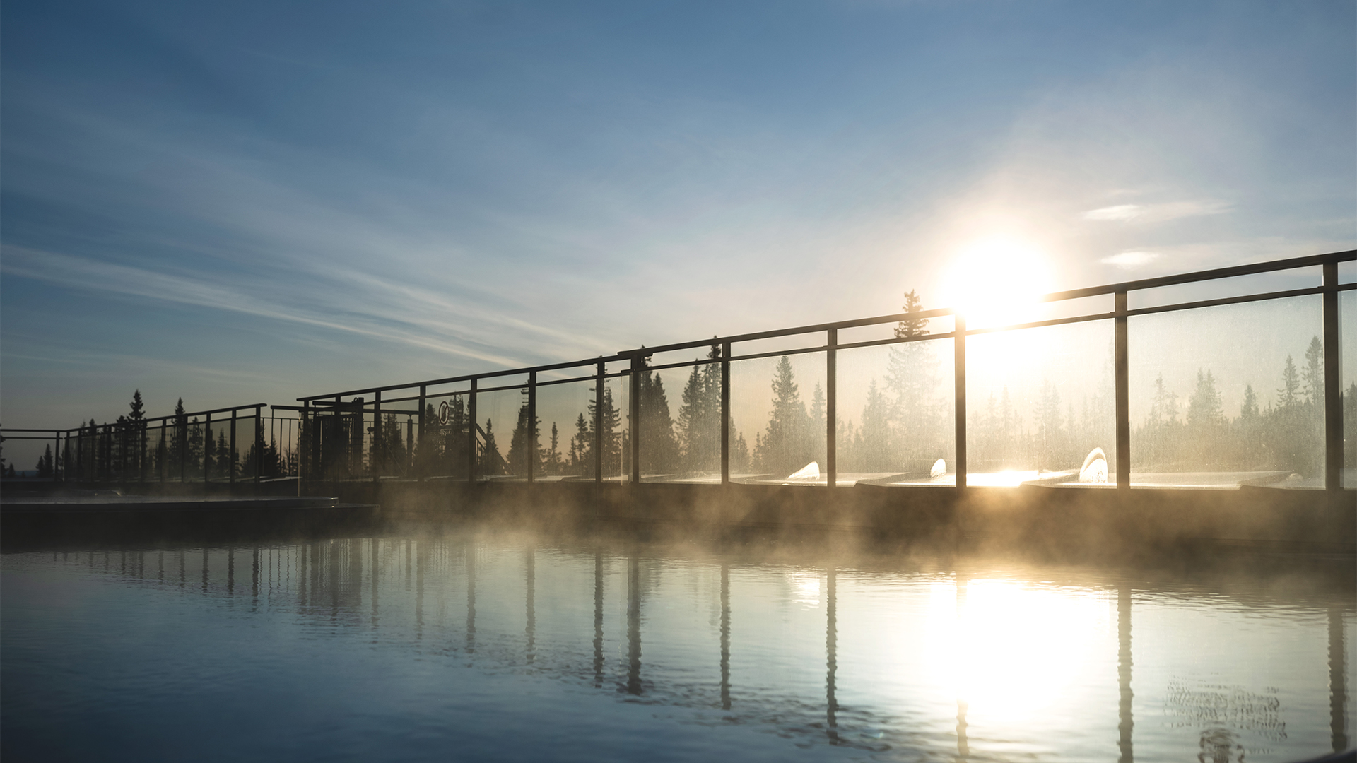 Soluppgång över en ångande utomhuspool med träd i bakgrunden, klarblå himmel