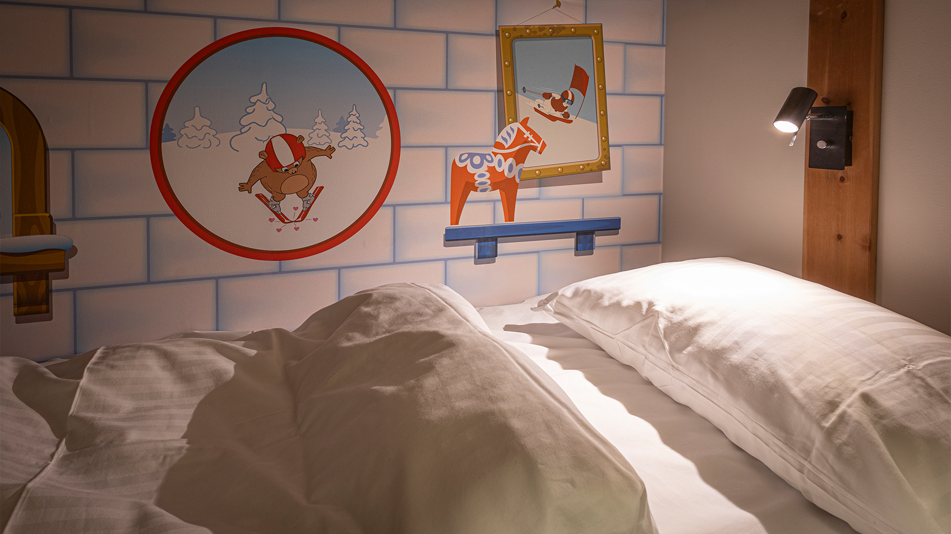 Bett mit weißer Bettwäsche in einem Raum, dekoriert mit Kinderzeichnungen von Zeichentricktieren, einschließlich eines ski fahrenden Bären und Rentiers, sowie Wandleuchten zur Beleuchtung
