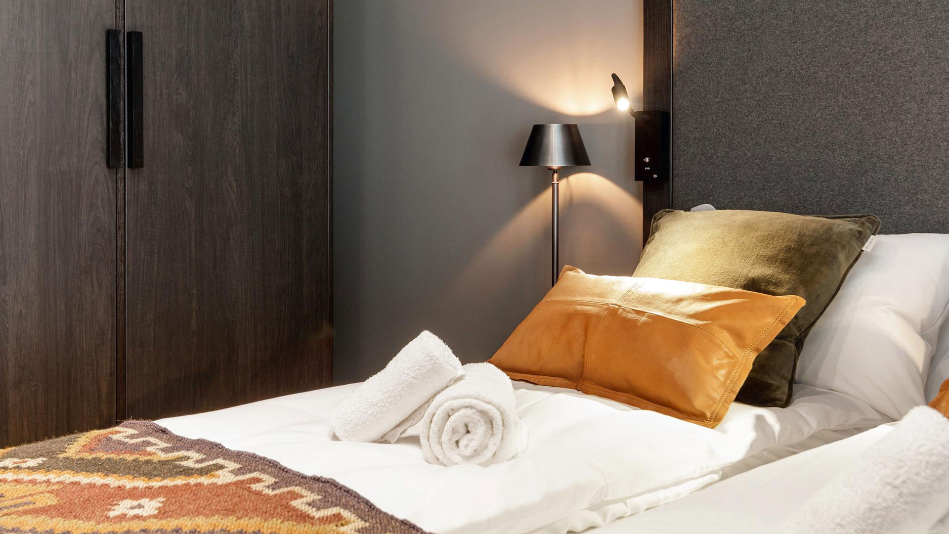 Rom med moderne innredning som viser en komfortabel seng med hvitt sengetøy, fargerike puter og et dekorativt teppe. En lampe og garderobe ses i bakgrunnen