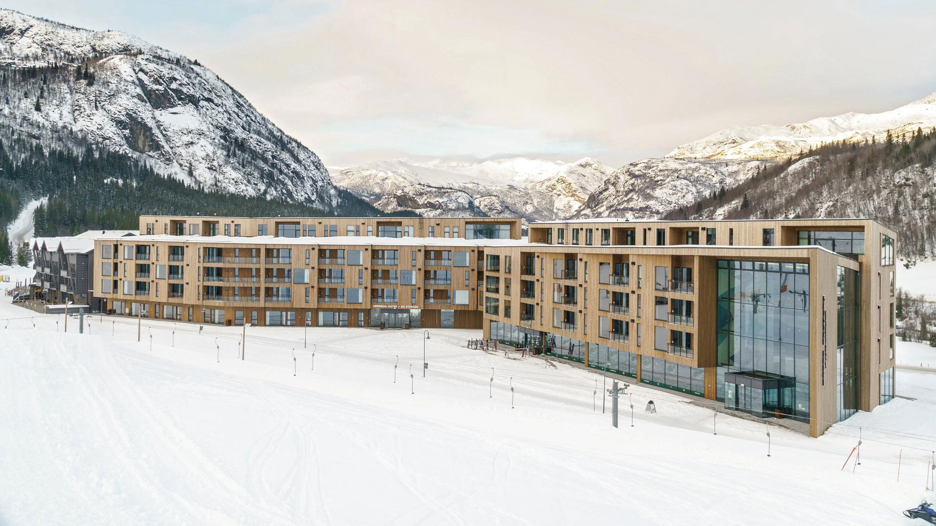 Modern fjällhotell med snöklädda fjäll och skidbackar i bakgrunden, trä- och glasfasad mot vintrig natur