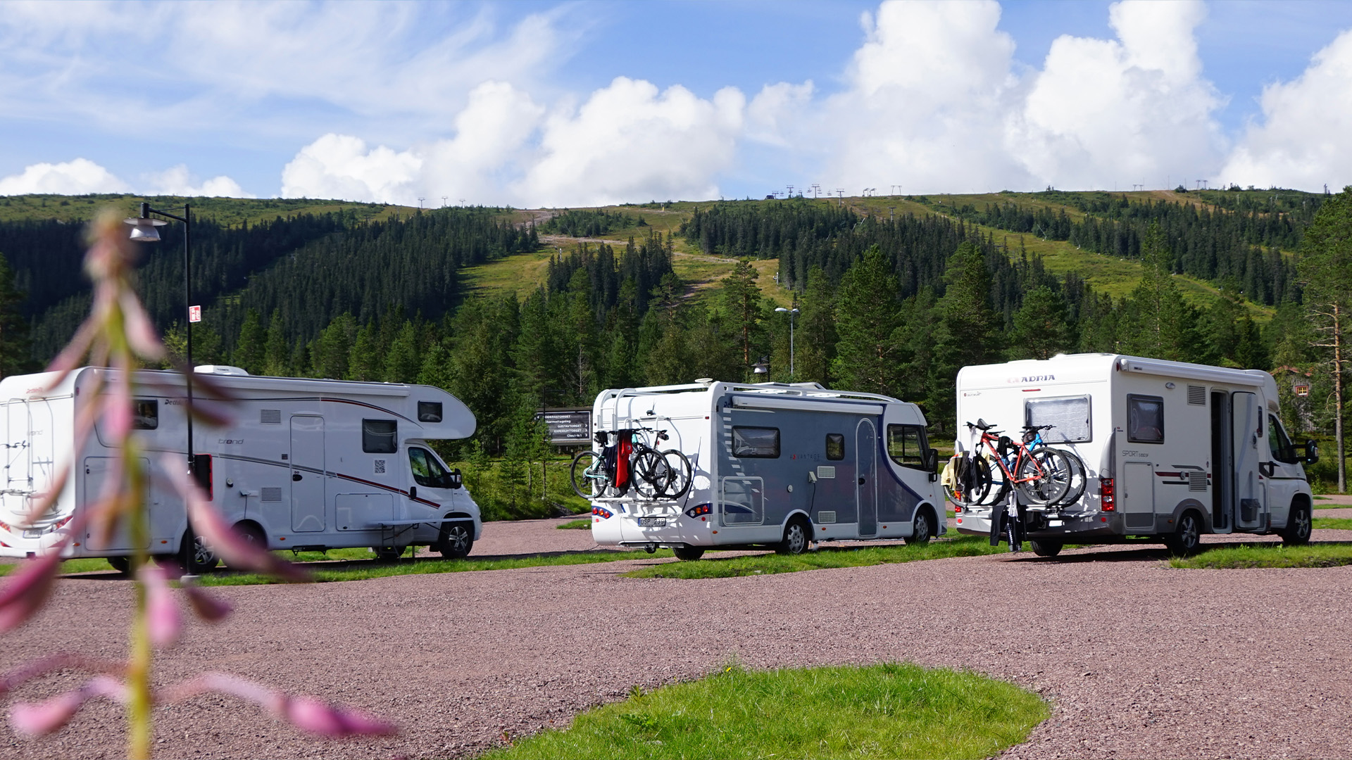 Husvagnar parkerade på en campingplats med cyklar monterade bakpå, omgivna av gröna skogar och berg under en blå himmel med moln