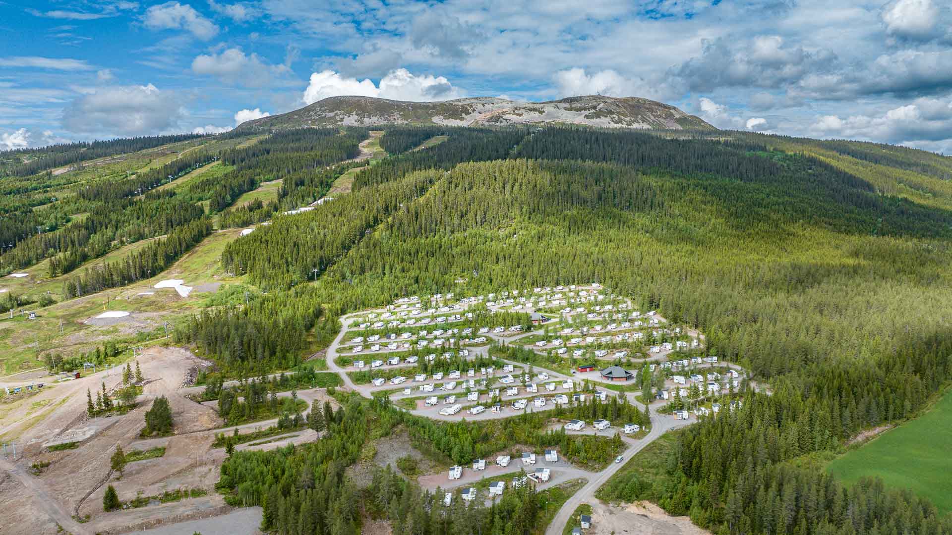 Vy över Bydalsfjällen med skogsklädda sluttningar och en campingplats full av husvagnar i förgrunden under en blå himmel med moln