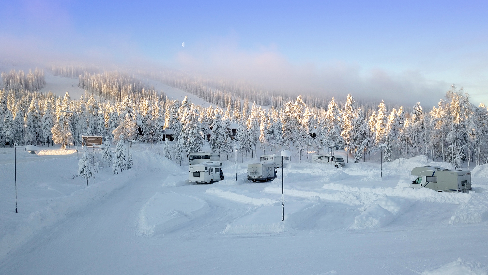 Snötäckt vinterlandskap med husbilar parkerade i ett campingområde, omgiven av frostiga tallar under en klar blå himmel