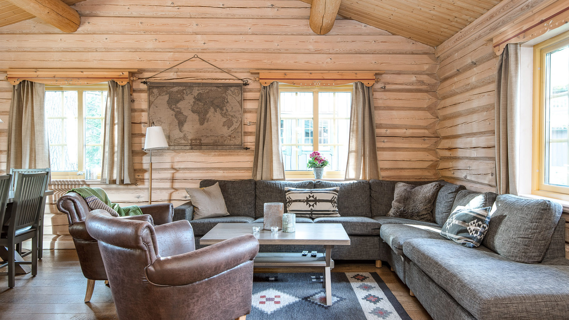 Rustikt stue i tømmerhytte med grå hjørnesofa, brune skinnlenestoler, verdenskart på veggen og vinduer med gardiner