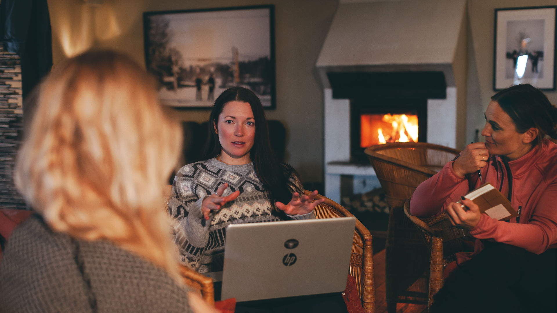 Tre personer har ett samtal i ett rum med öppen spis. En kvinna håller en laptop och pratar med de andra, som lyssnar och tar anteckningar. Rummet är mysigt och varmt upplyst
