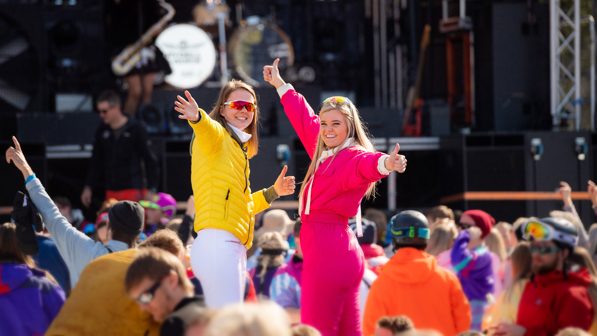 To personer i fargerike vinterklær viser tommelen opp og ser glade ut på en utendørskonsert i løpet av dagen