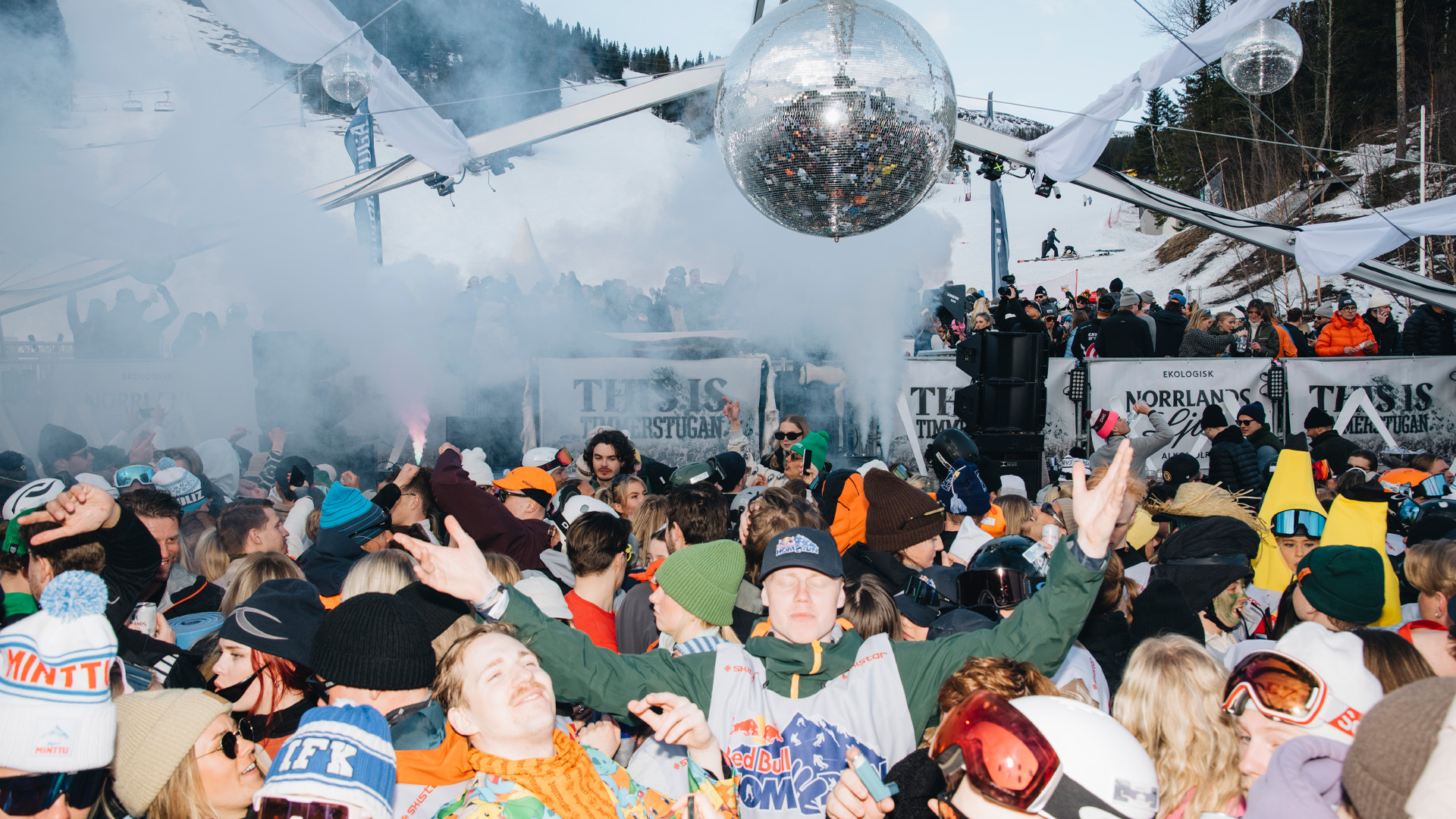 Unge mennesker fejrer på et skisportssted med en stor diskokugle og musik i baggrunden. Sne og vinterlandskab ses rundt omkring, hvilket skaber en livlig og festlig atmosfære