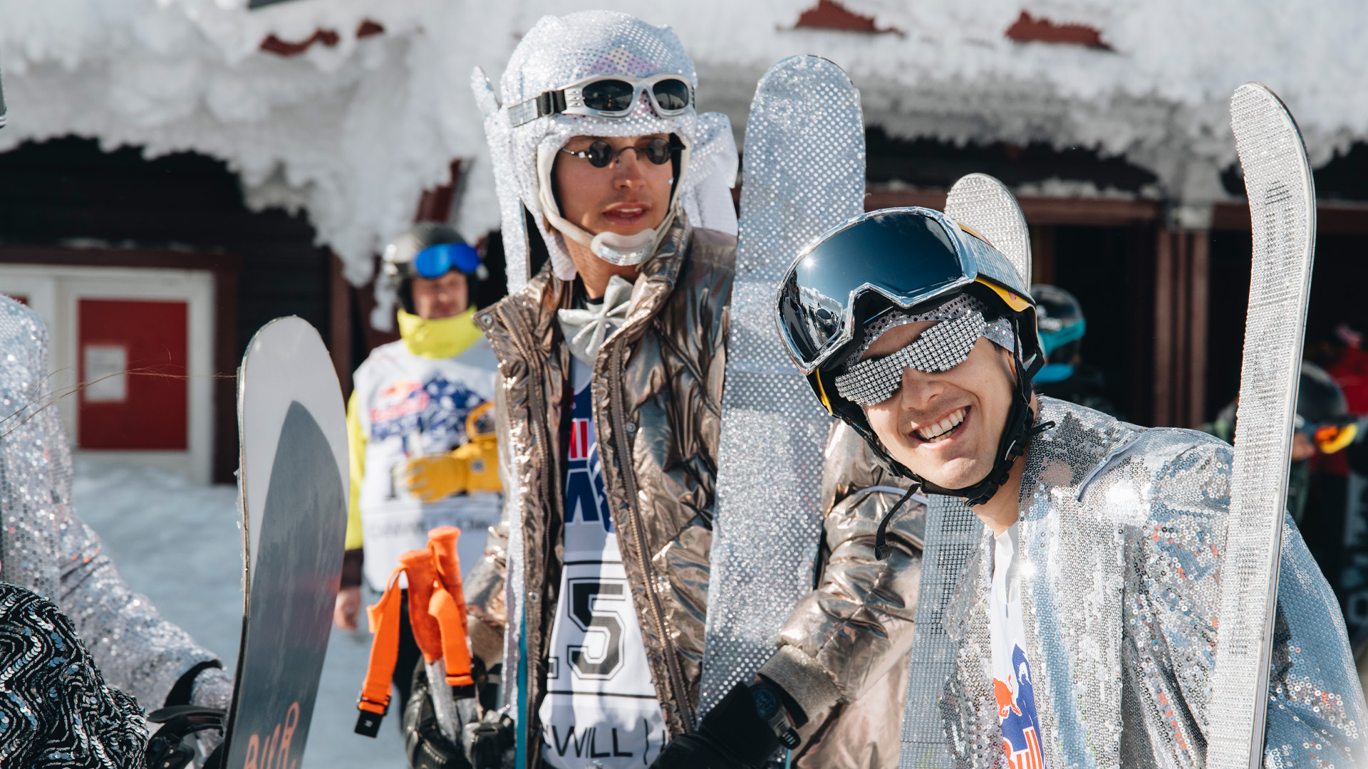 Personer klædt i glitrende sølvdragter og solbriller holder ski og snowboard, smilende og klar til snesport, med en sneklædt hytte i baggrunden