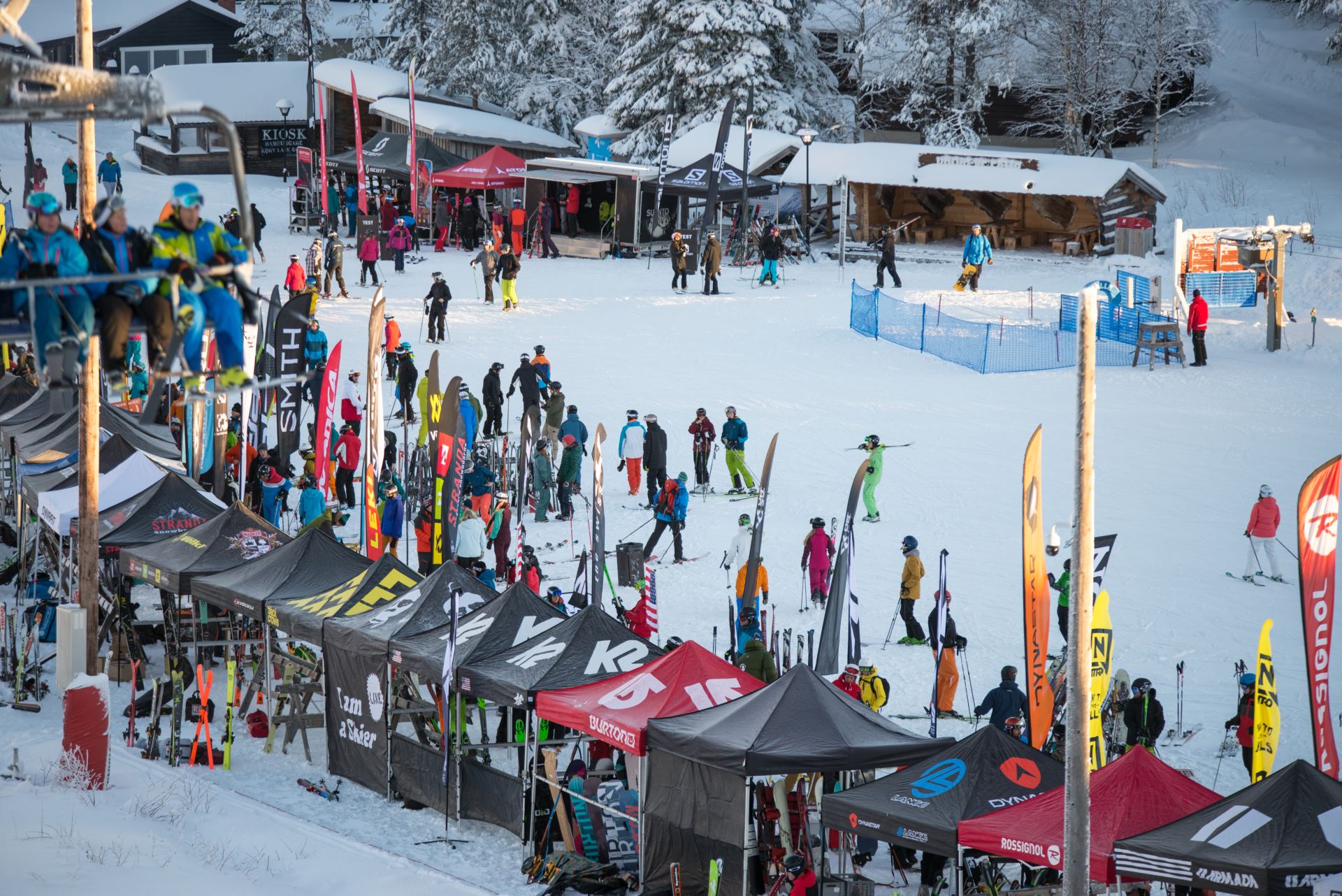 Skidåkare samlas vid en skidort med färgglada tält och flaggor, snötäckta träd i bakgrunden och människor på en lift i förgrunden
