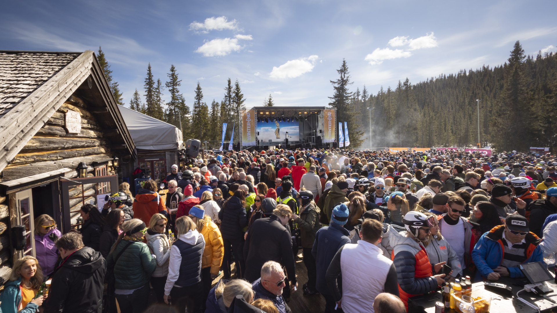 Stor folkmassa vid utomhusscen under vårfestival i fjällmiljö, med soligt väder och omgivande tallskogar